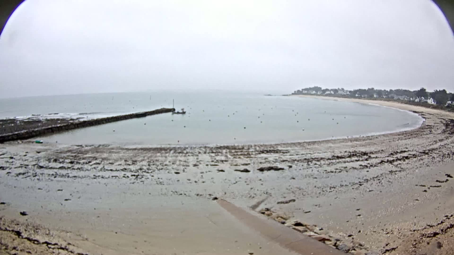 A wide, mostly empty sandy beach curves around a calm bay under an overcast sky, with a small pier extending into the water.