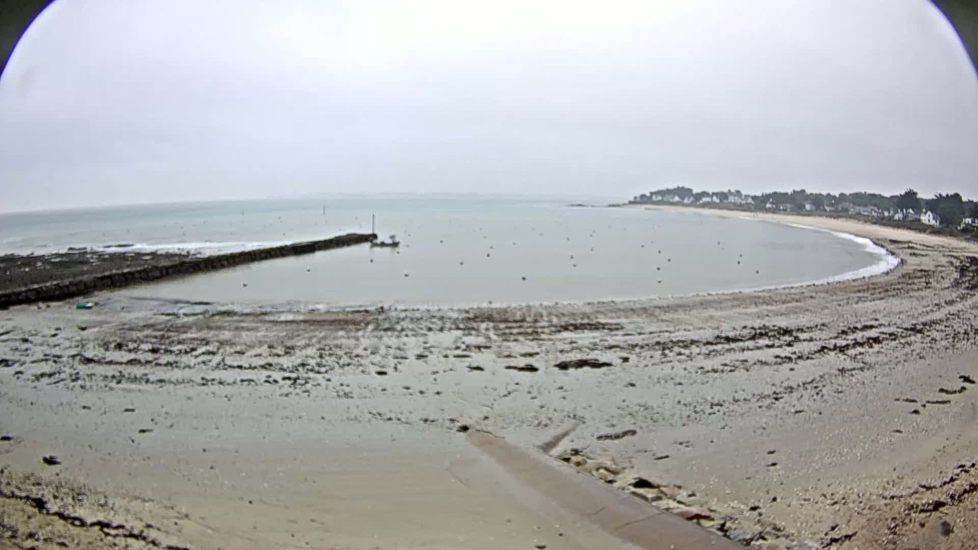 A mostly cloudy day reveals a small, calm bay with a muddy shoreline, a short stone jetty, and a few small boats.