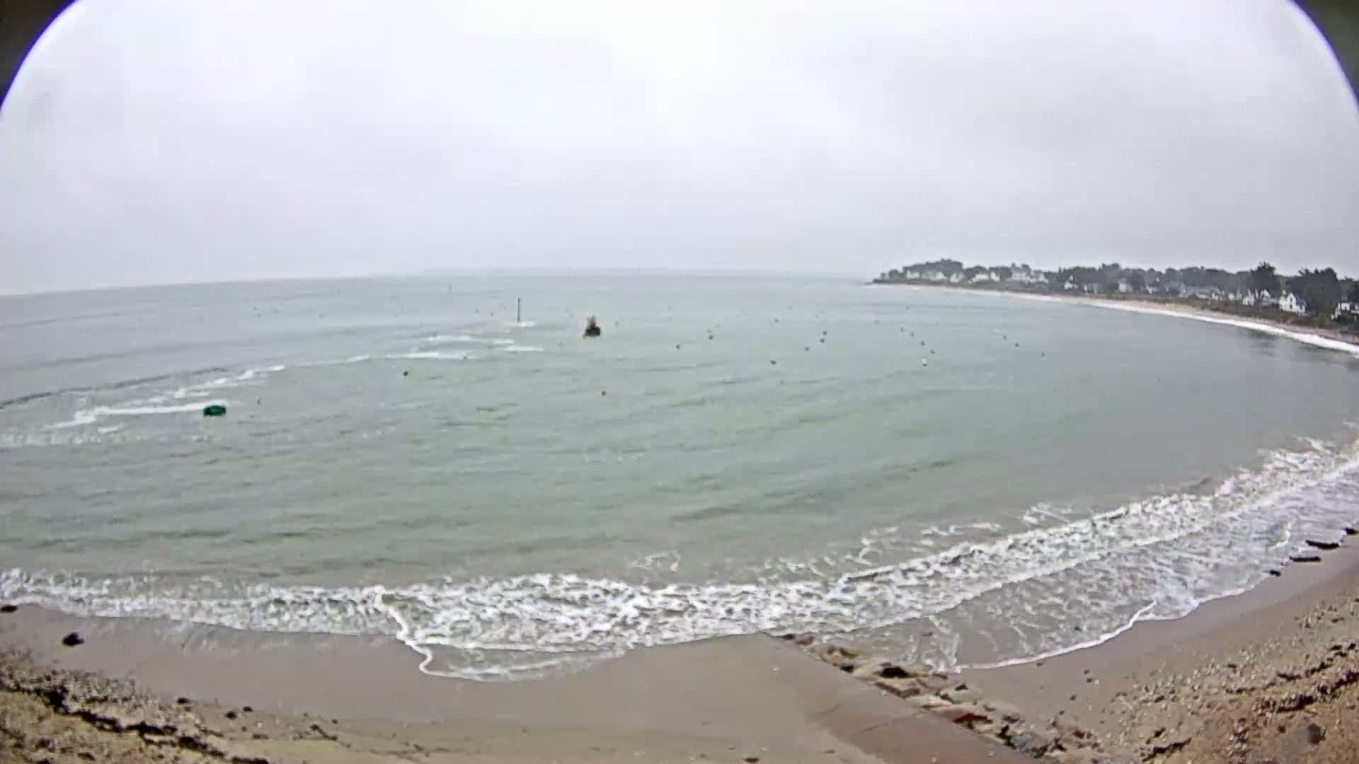 A sandy beach with small waves laps the shore of a calm bay under an overcast sky.