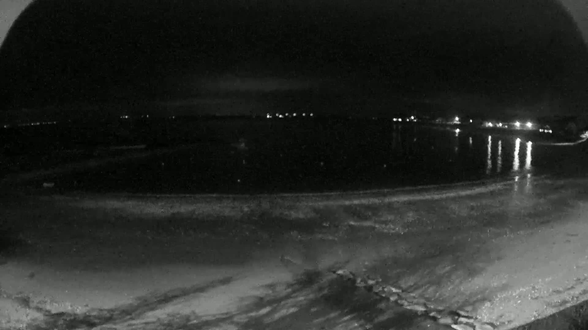 A dark beach at night is shown with calm water reflecting distant lights.