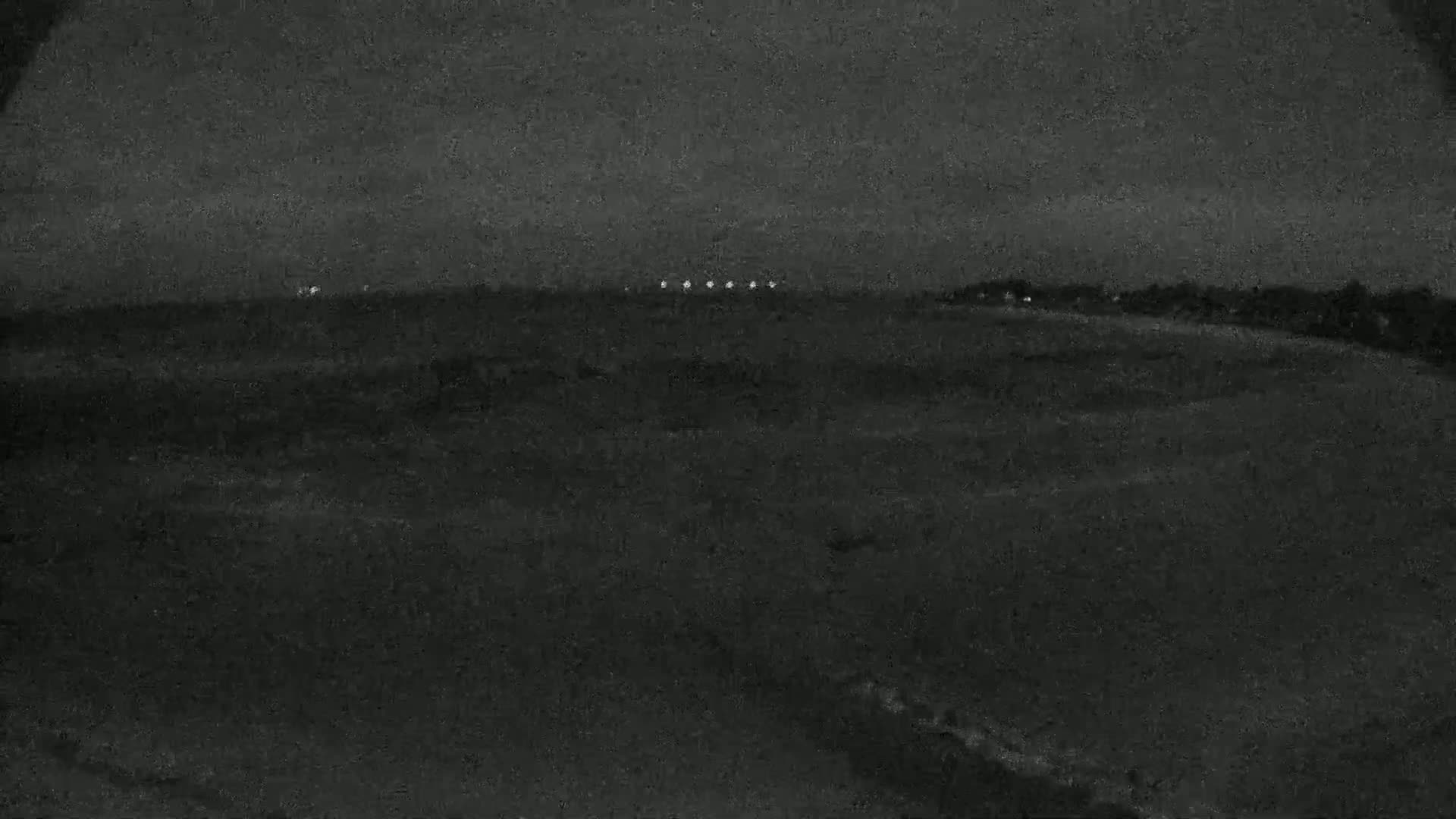 A dark, nighttime landscape shows a flat expanse of land with a few distant lights on the horizon under a mostly clear sky.