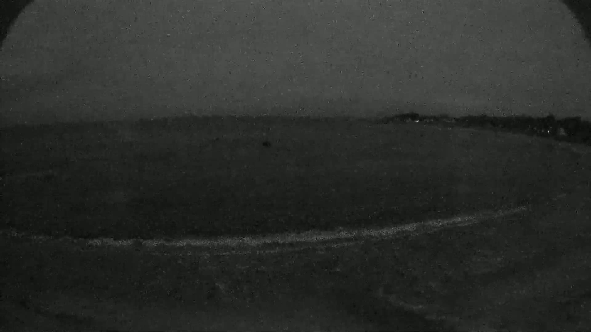 A dark, possibly nighttime, outdoor scene shows a body of water with a faint line of white along the bottom, possibly waves, and a dark landmass in the background under a dark, possibly rainy, sky.