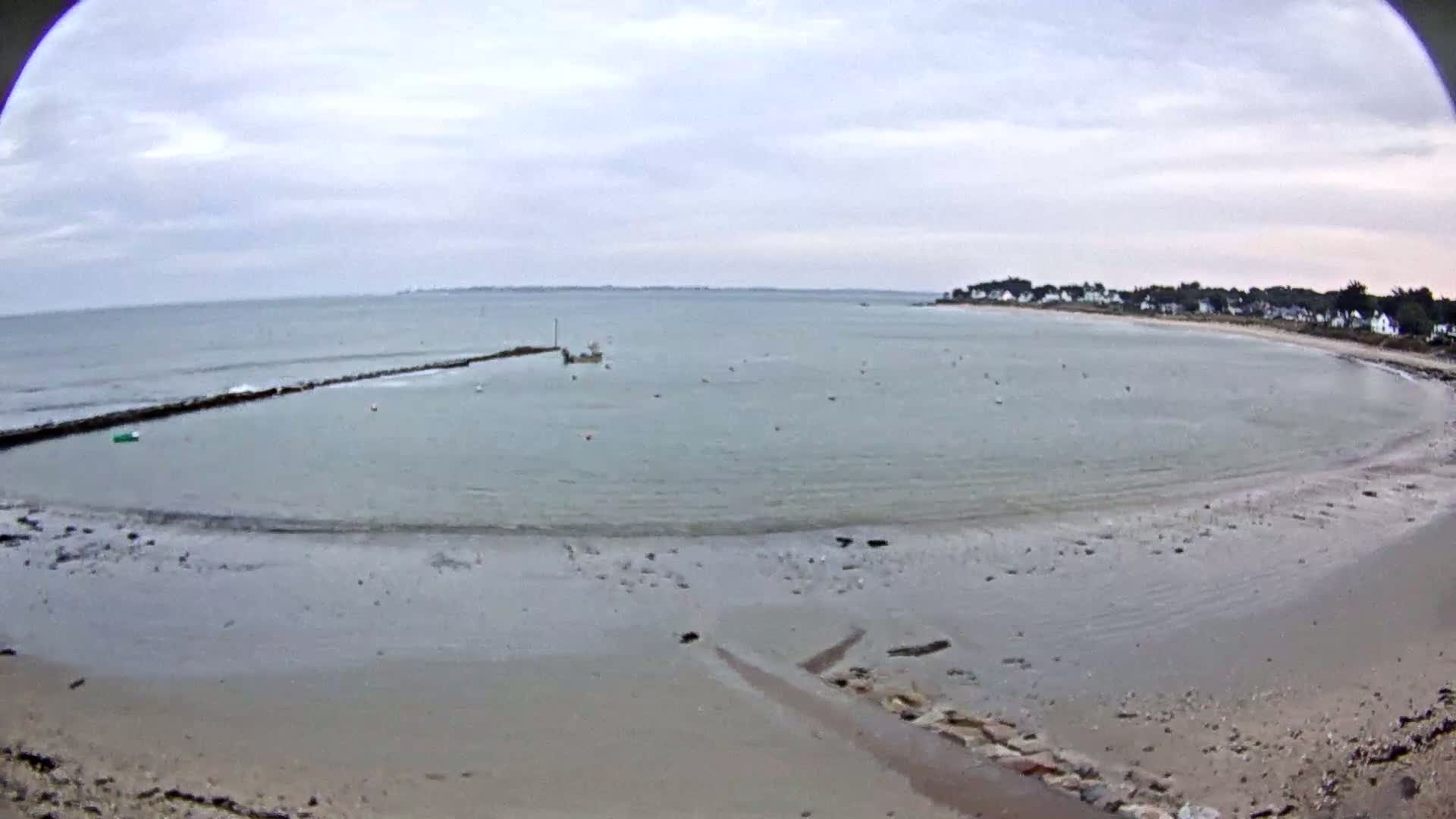 A mostly calm bay with a small boat near a stone jetty, under an overcast sky, reveals a sandy shore with a small village visible in the distance.