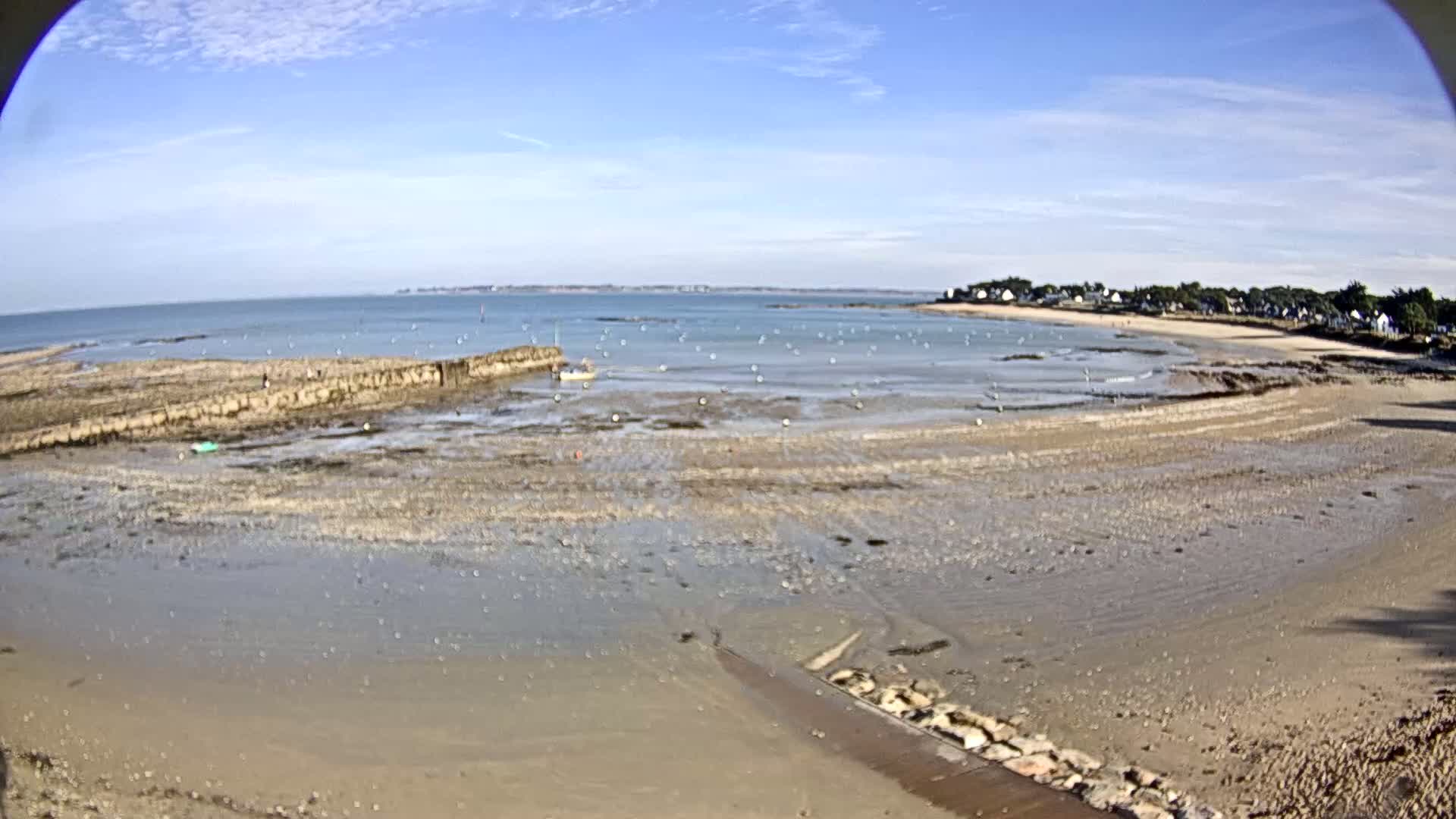 A mostly sunny day reveals a sandy, low-tide beach and calm bay with a small boat and several markers in the water, backed by a line of trees and buildings.