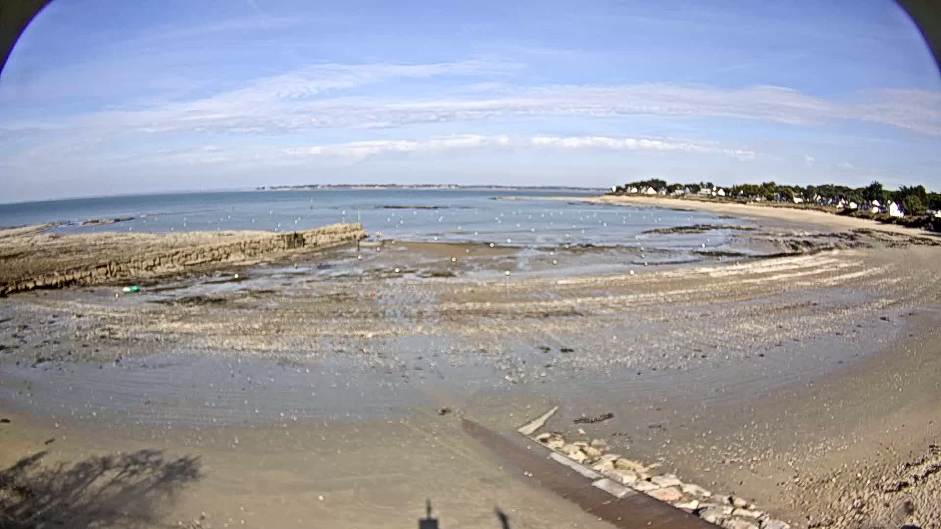 A mostly sunny day at a sandy beach with calm water, showing exposed mudflats at low tide and a small village in the distance.