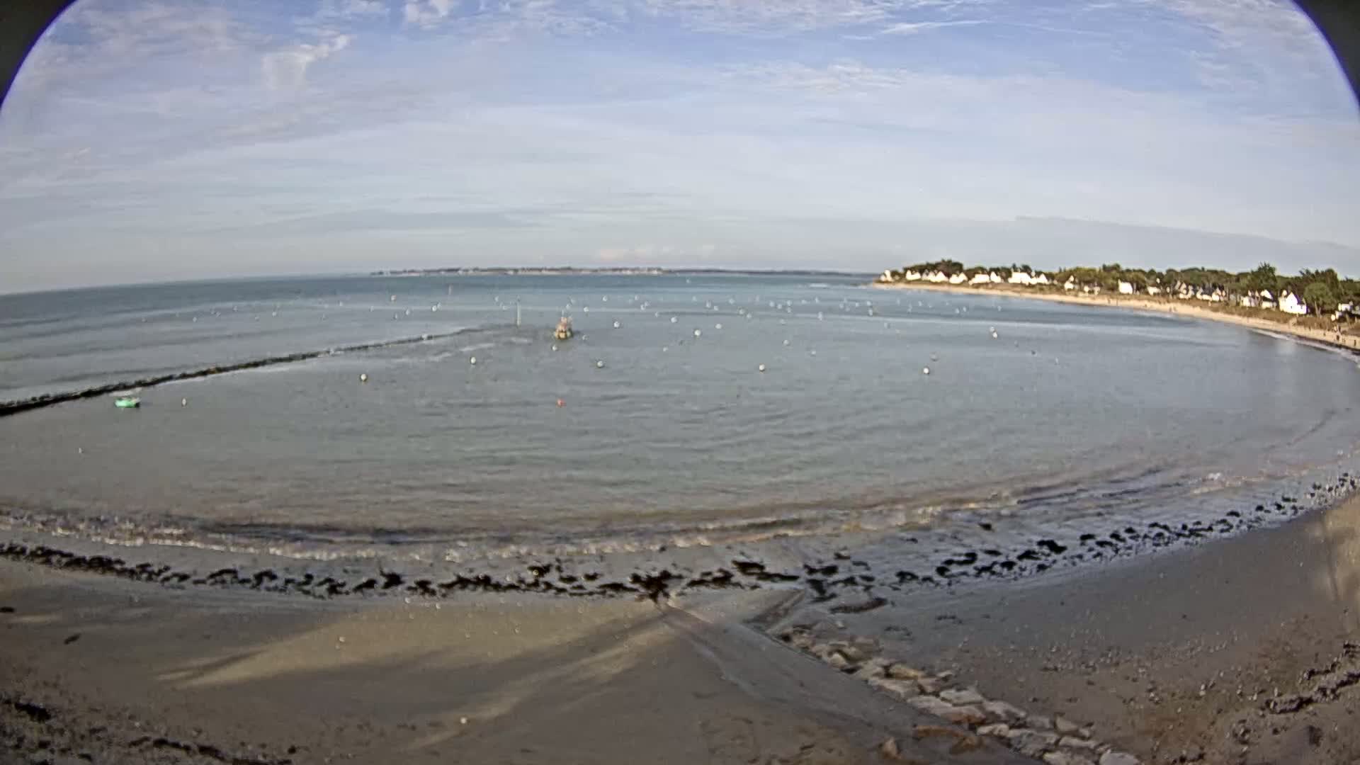A sandy beach curves around a calm bay under a mostly clear sky, with a few small boats and buoys visible in the water.