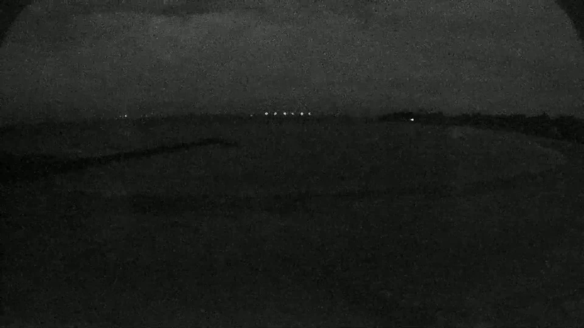 A dark, nighttime landscape with a few distant lights visible under a cloudy sky.