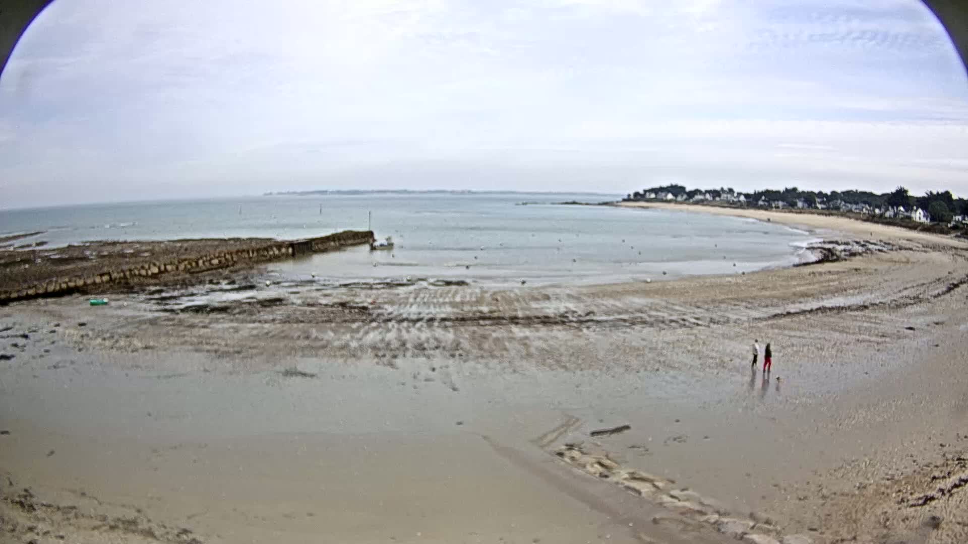 A wide shot shows a mostly flat, sandy beach at low tide with two people walking near the water's edge, a calm bay, and a distant shoreline under a cloudy sky.
