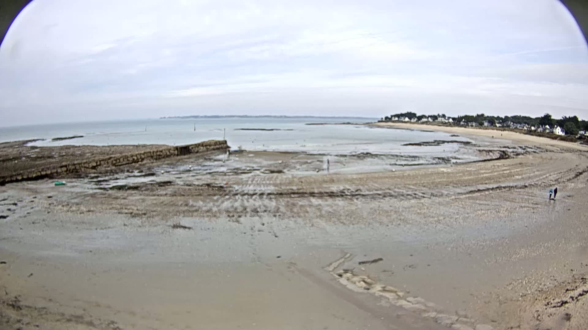 A wide, sandy beach stretches out to a calm, light-grey sea under a mostly cloudy sky.