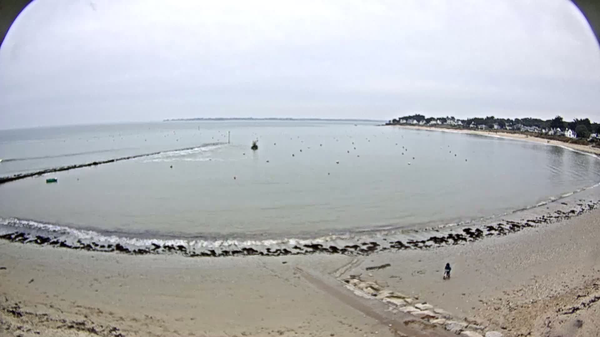 A calm, overcast day at a sandy beach with a small body of water partially enclosed by a rock jetty, showing a few small waves lapping the shore and a lone person walking on the beach.