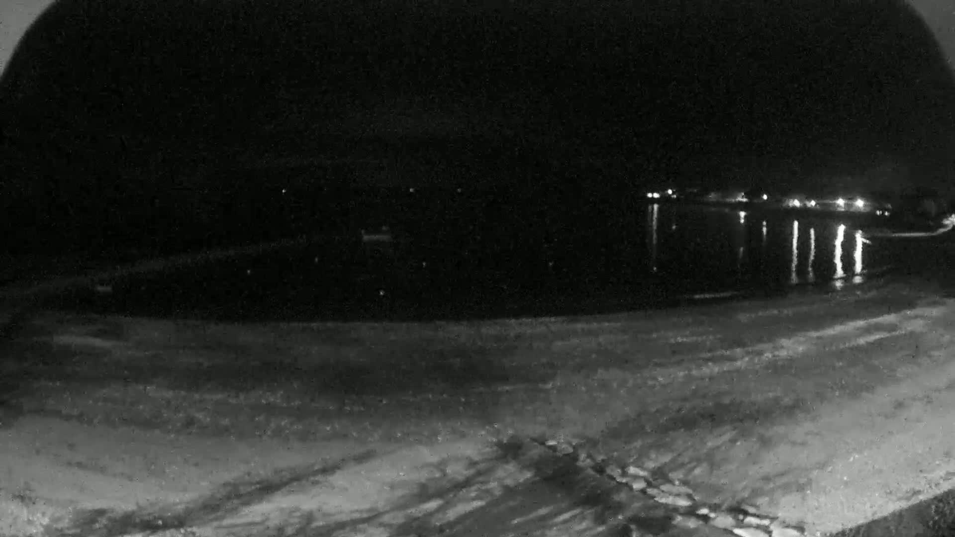 A nighttime, low-light view shows a dark body of water reflecting distant lights, with a sandy shoreline in the foreground.