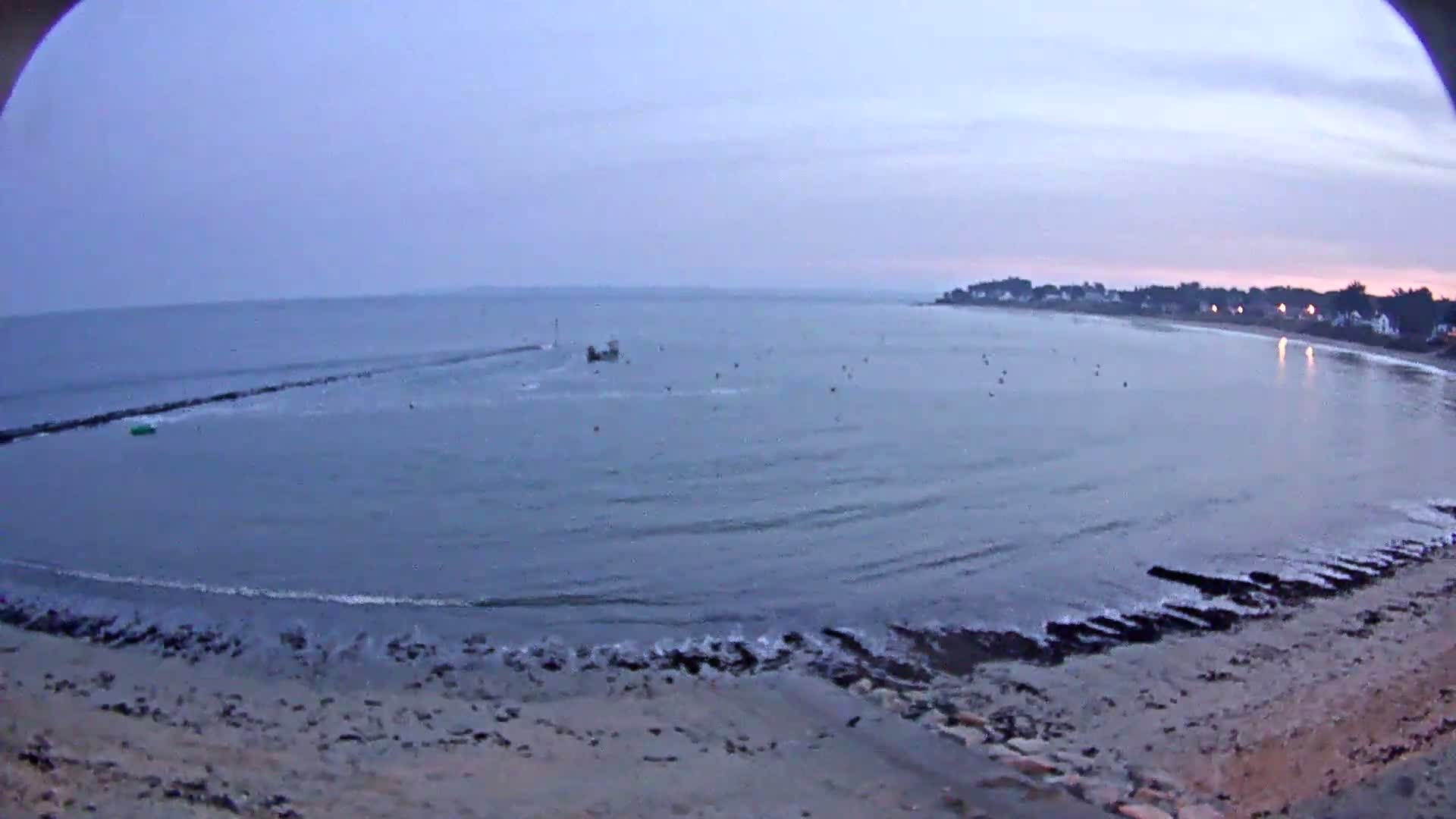 A calm, twilight sea laps a sandy shore near a distant town under a pale, cloudy sky.