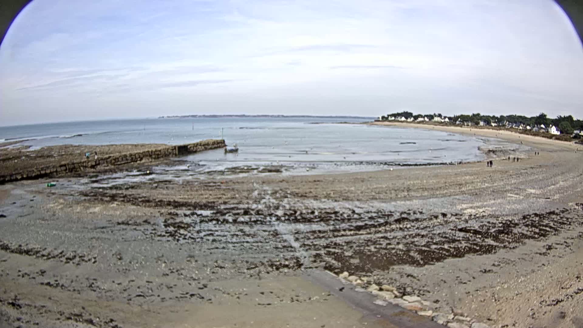 A mostly sunny day reveals a wide, sandy beach at low tide, with a small town visible in the distance, a low stone wall on the left, and a few people walking along the shoreline.
