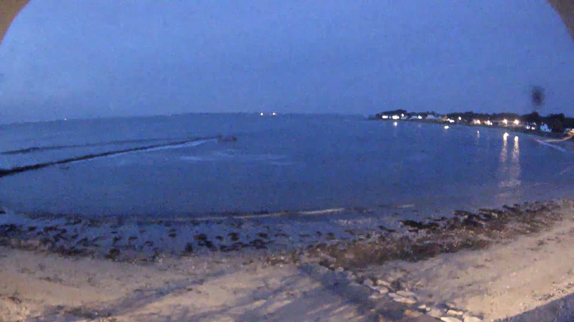 A sandy beach at dusk, under a dark blue sky, meets a calm body of water with a distant shoreline featuring illuminated buildings.