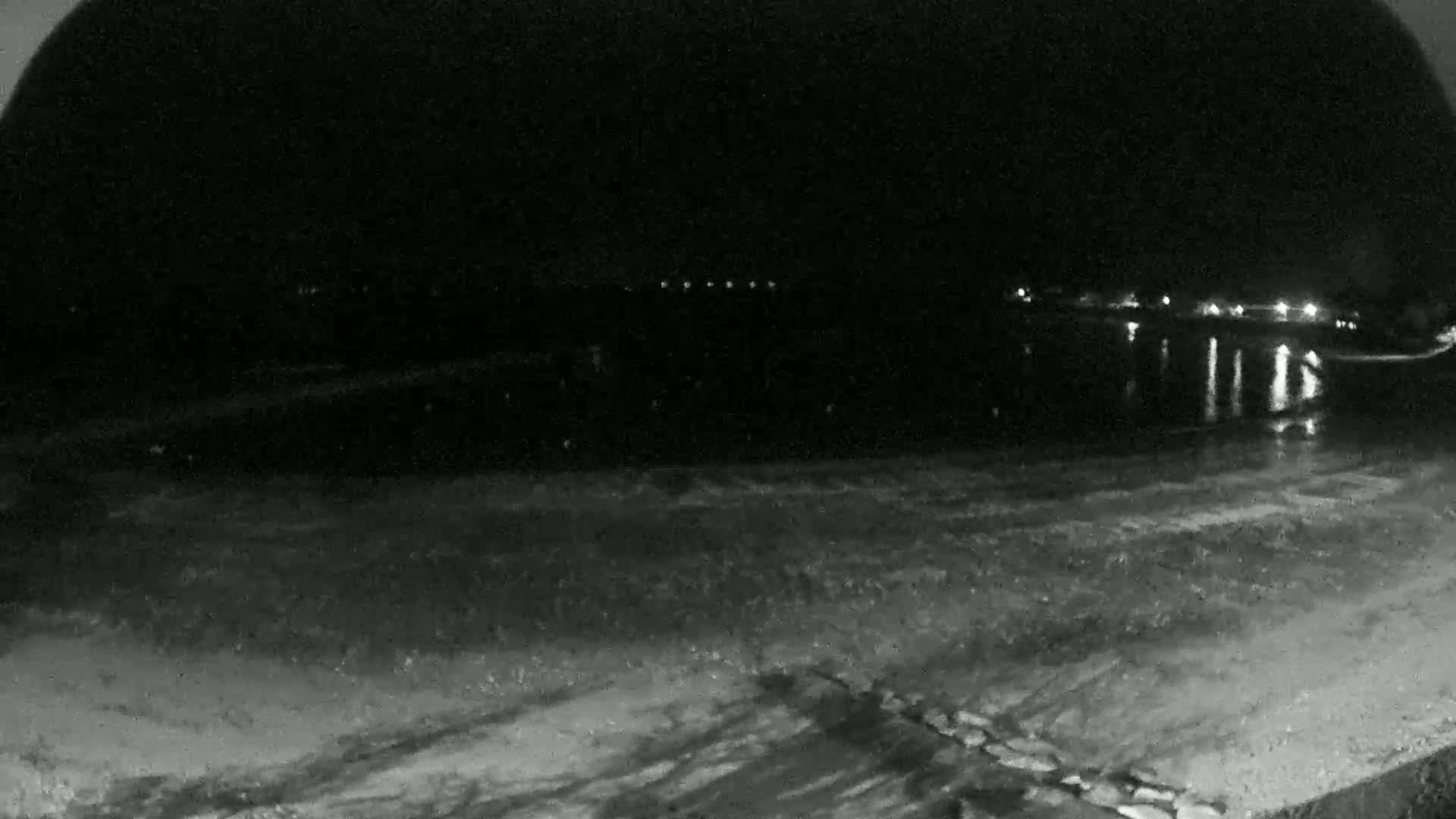 A nighttime, low-light view of a dark body of water reflecting distant lights, with a foreground of textured, possibly icy ground.