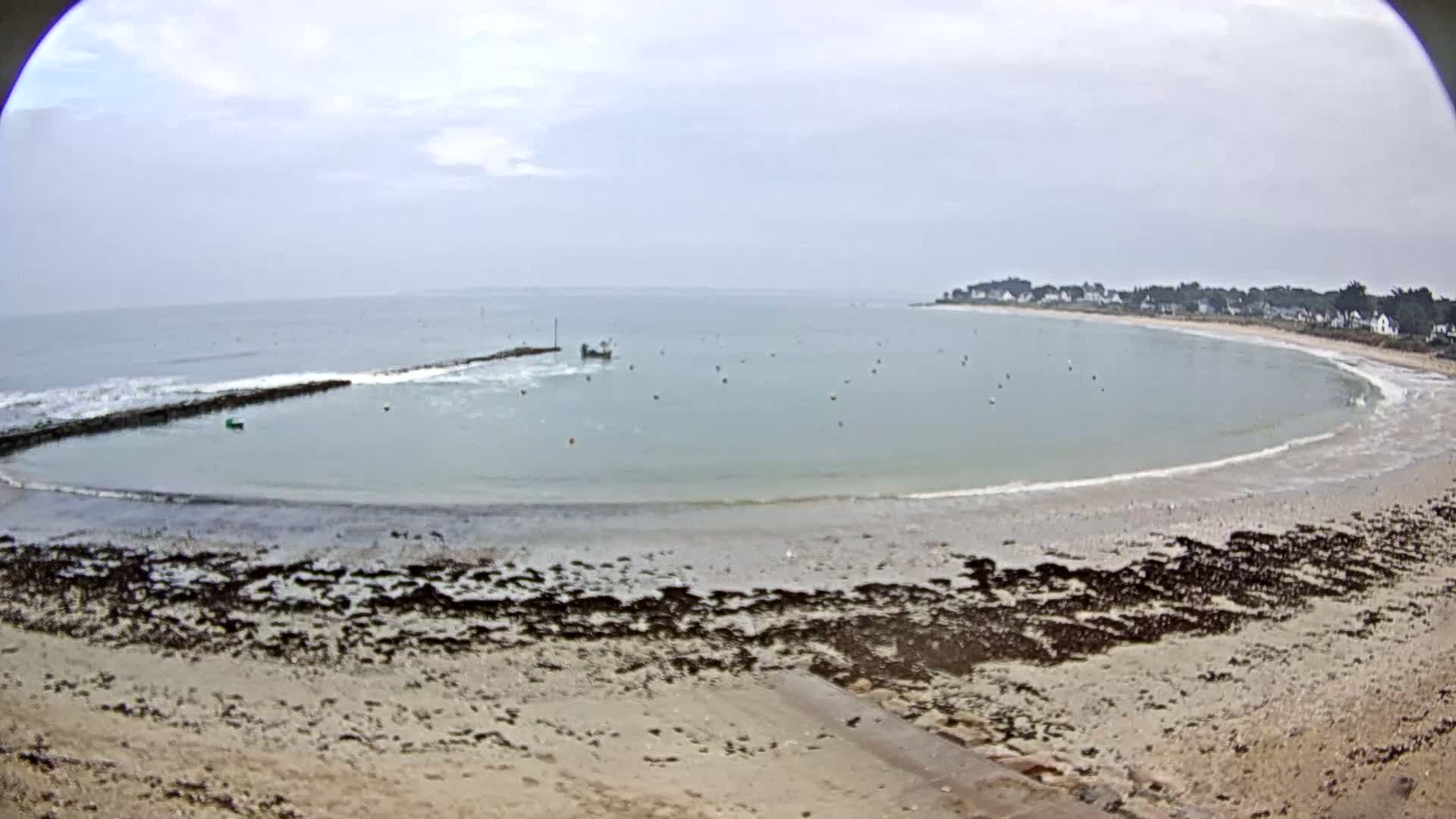 A sandy beach curves around a calm, light-green bay under an overcast sky, with a small boat visible in the water and a breakwater partially visible near the shore.