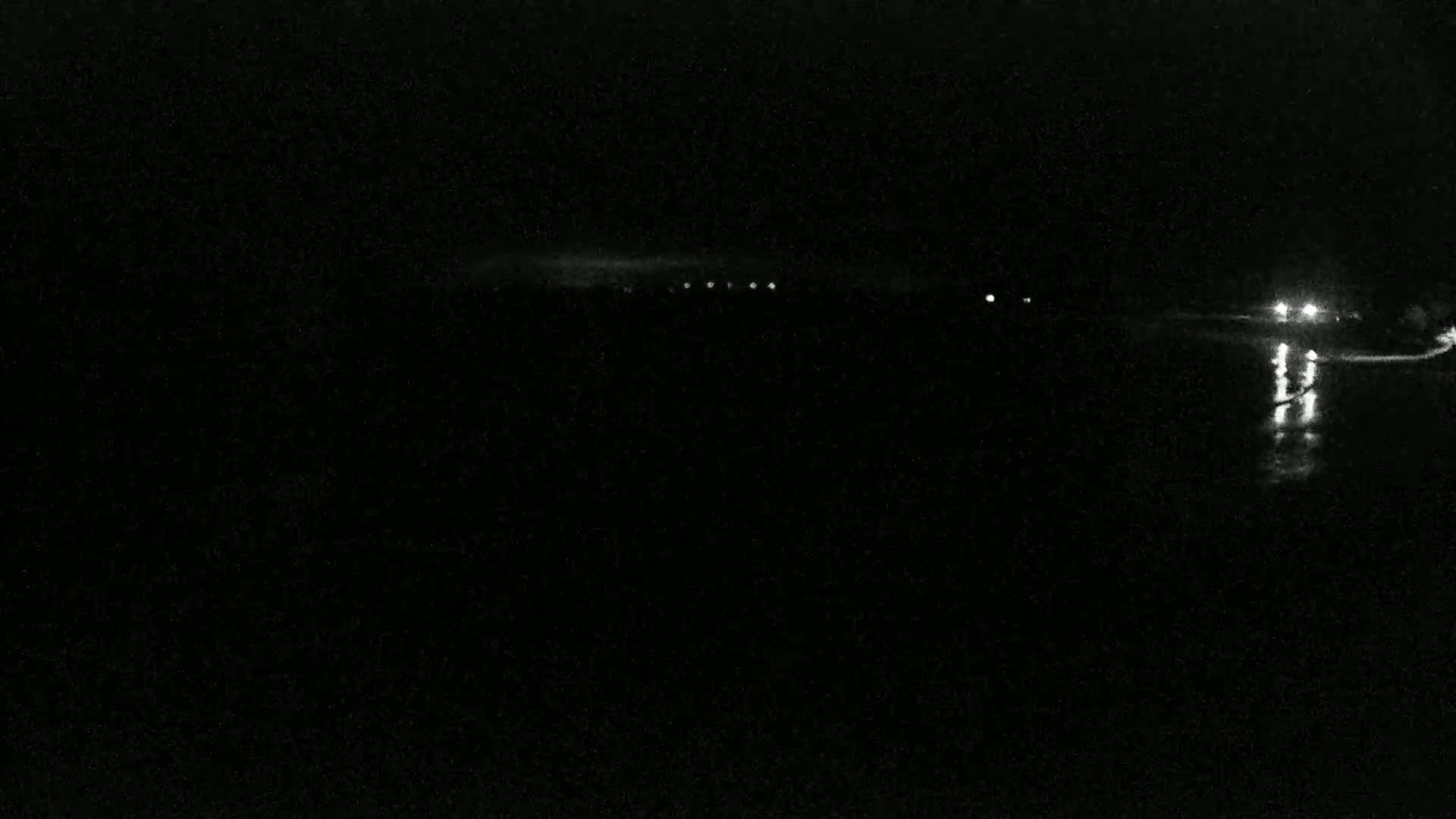 A nighttime, dark scene shows a line of distant lights reflected on dark water.