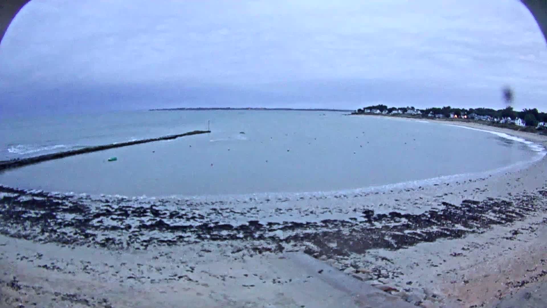 A calm, overcast day reveals a sandy beach curving around a bay, with a low-lying breakwater and a distant shoreline dotted with houses.