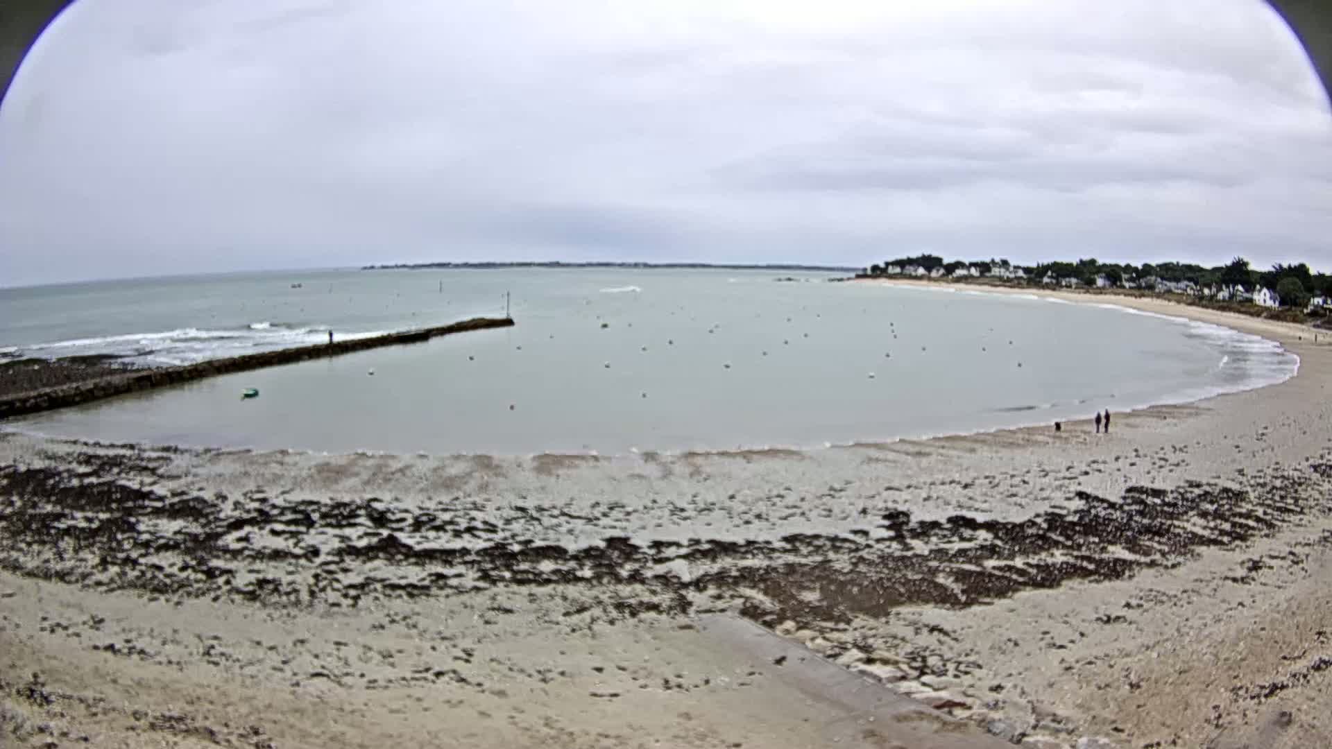 A sandy beach curves around a calm bay under an overcast sky, with a small stone jetty and a few people visible in the distance.