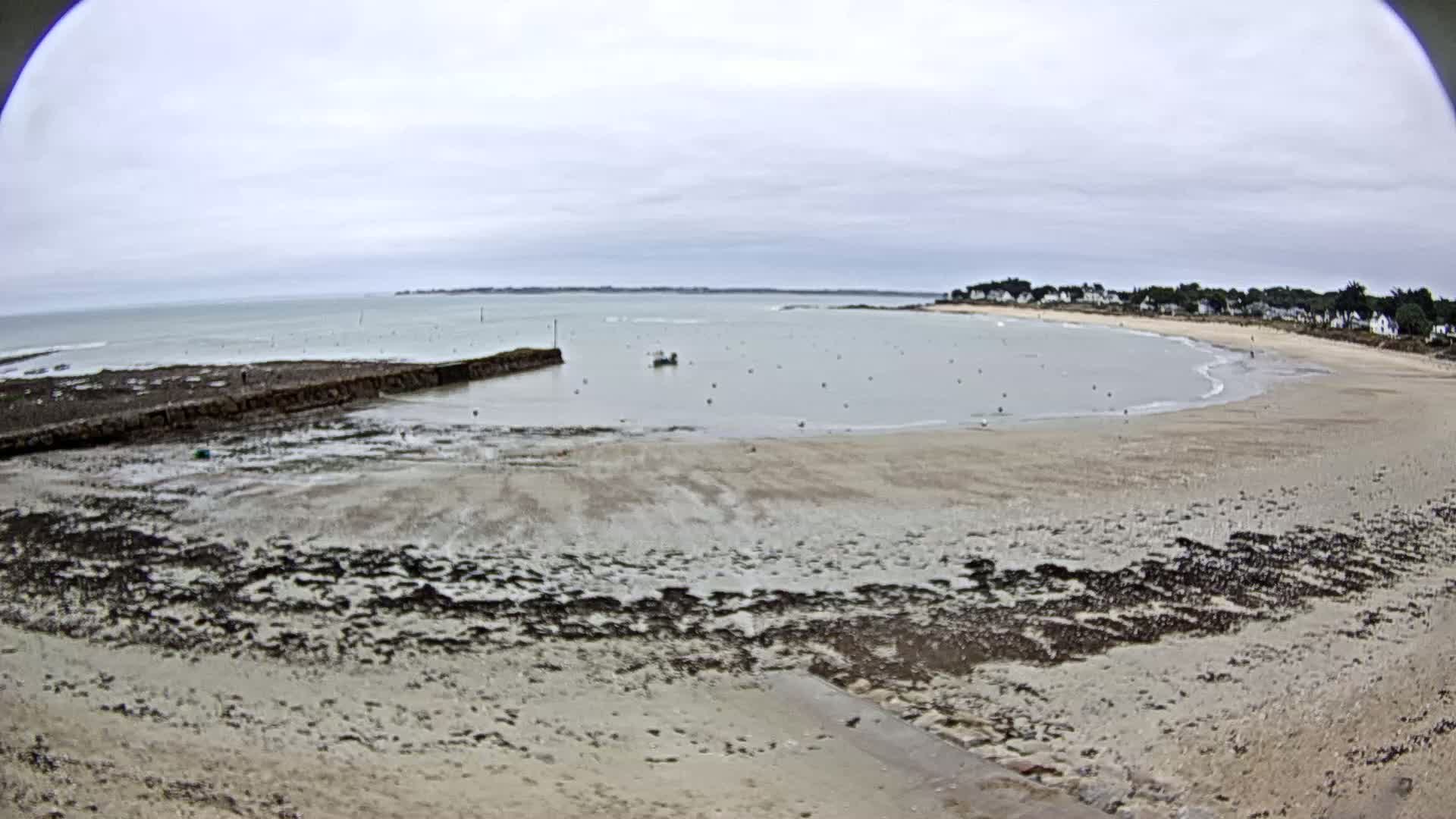 A sandy beach and calm bay are visible under an overcast sky, with a small stone pier and a few boats in the water.