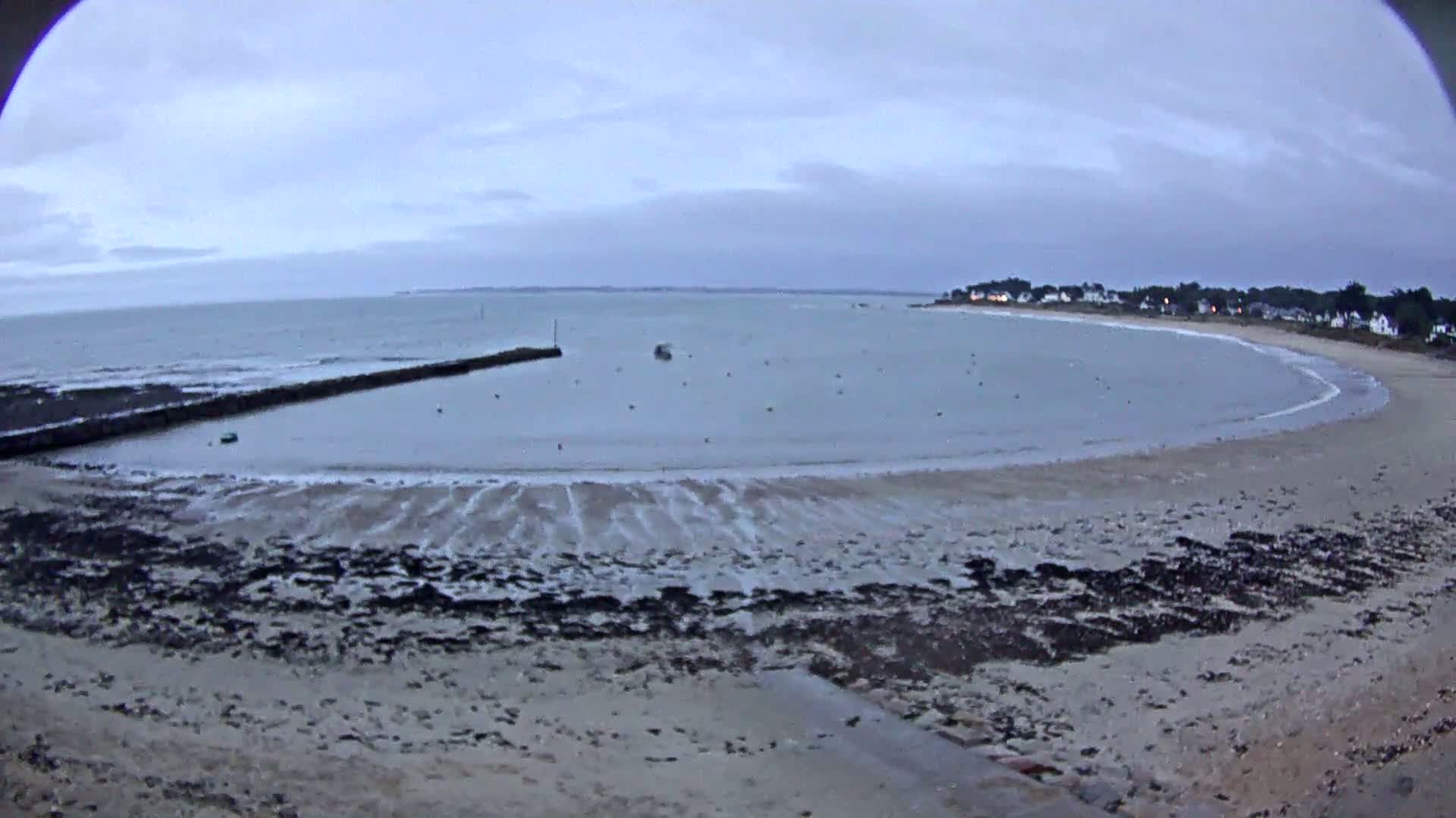 A calm, overcast day at a sandy bay with a small harbor, featuring a low-lying breakwater and a few boats in the water.