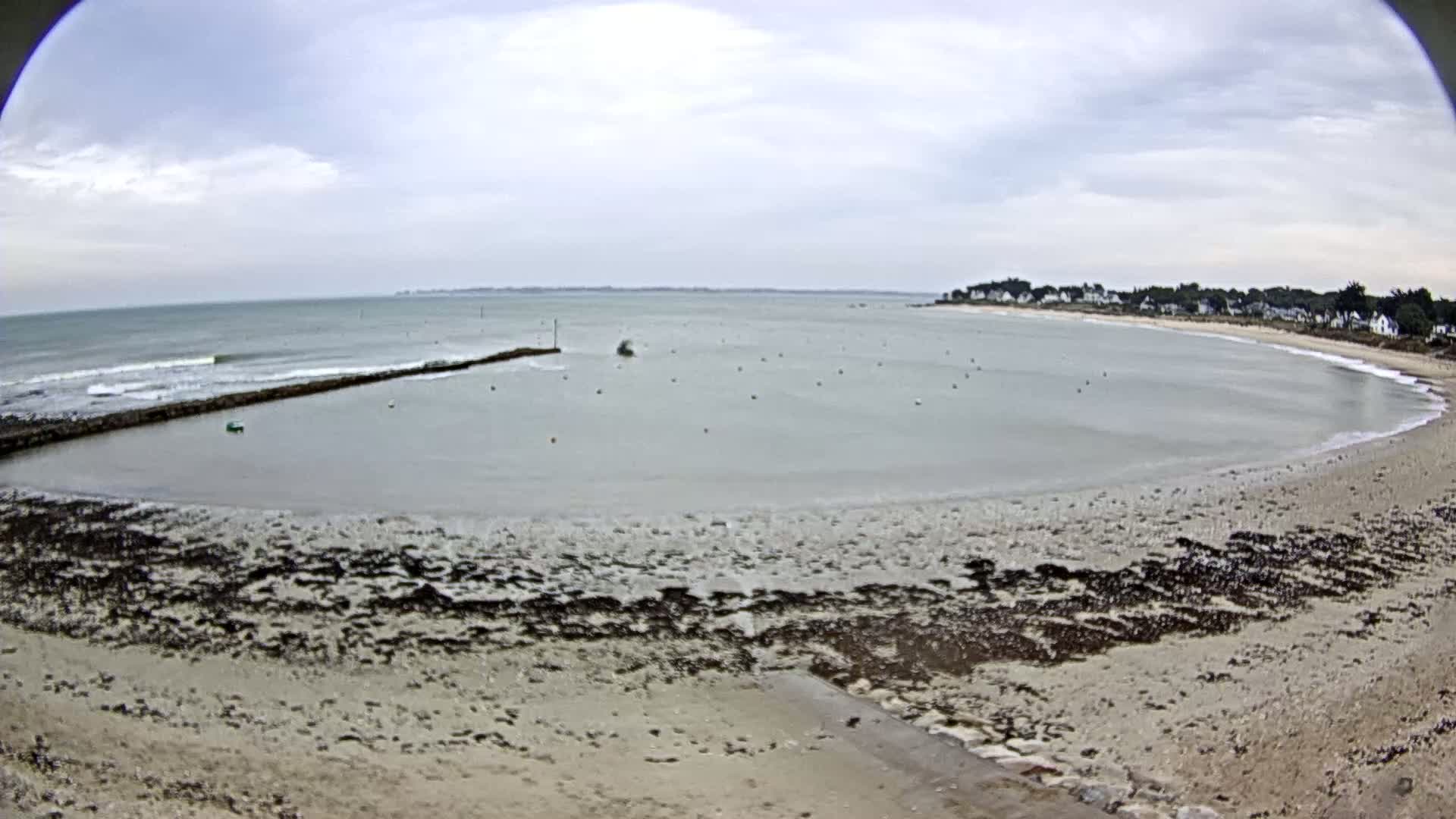 A calm, overcast day shows a sandy beach curving around a bay with small waves breaking against a low wall, and a small town in the distance.