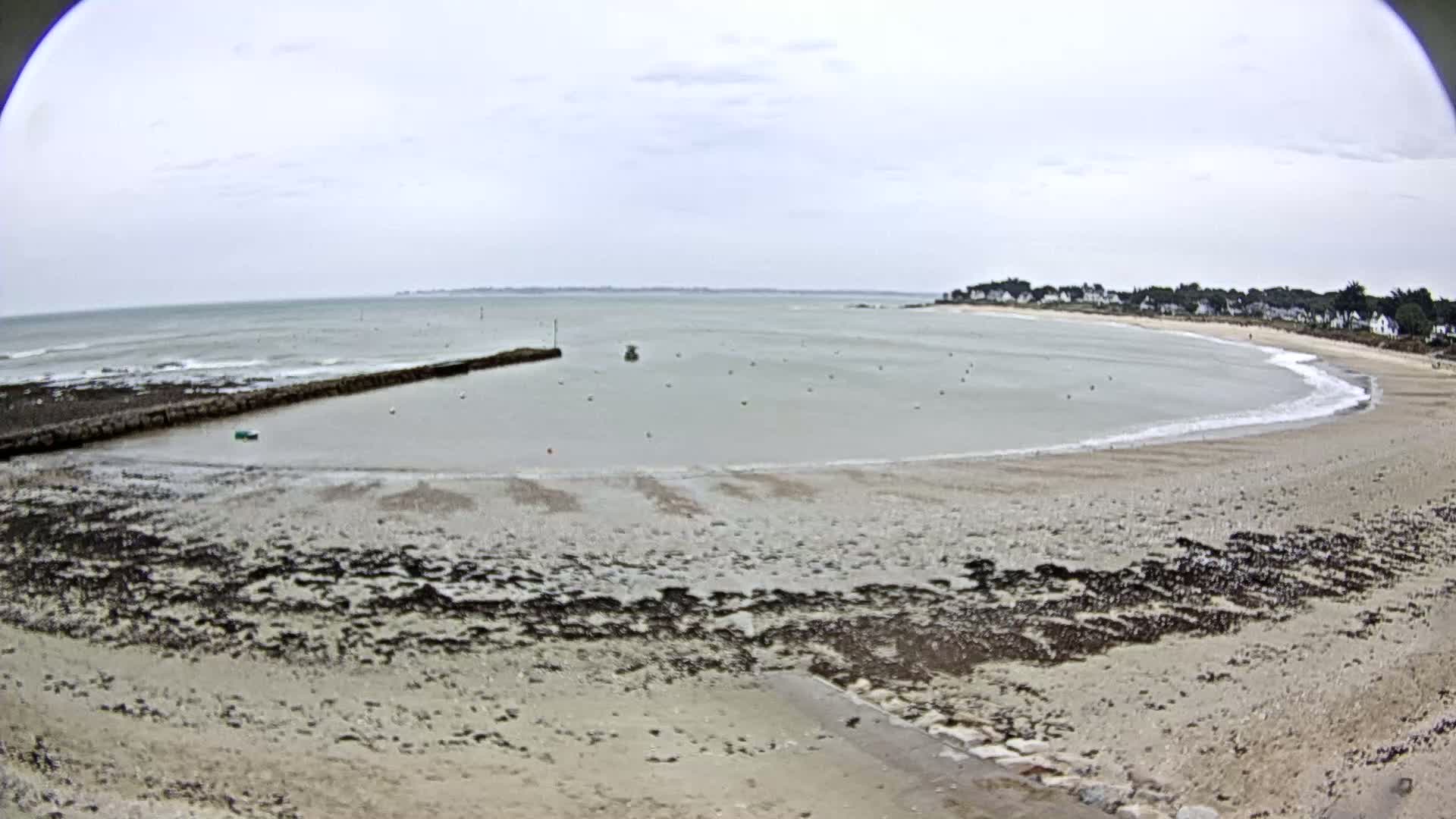 A sandy beach curves around a calm bay under an overcast sky, with a small, man-made harbor visible at the left.
