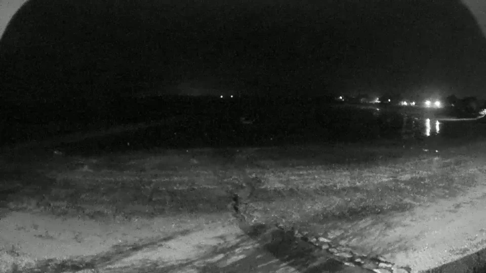 A nighttime, low-light view shows a dark body of water reflecting distant lights, with a snow-covered foreground extending to the bottom of the frame.