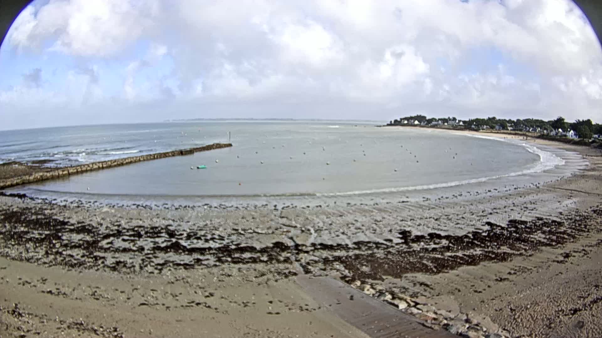 A mostly cloudy day reveals a sandy beach curving around calm, shallow water, with a stone jetty partially enclosing a small harbor.