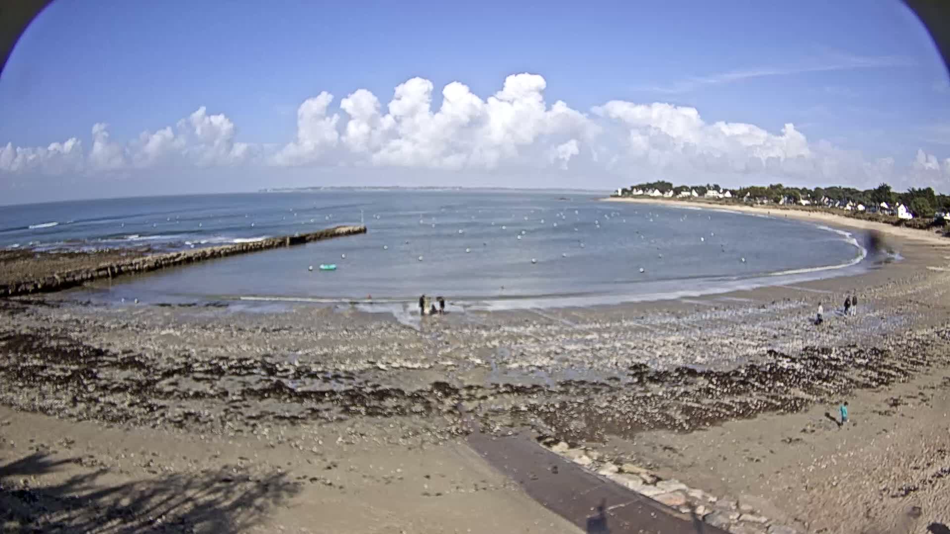 A sandy beach with a calm bay and a few people, under a partly cloudy blue sky.