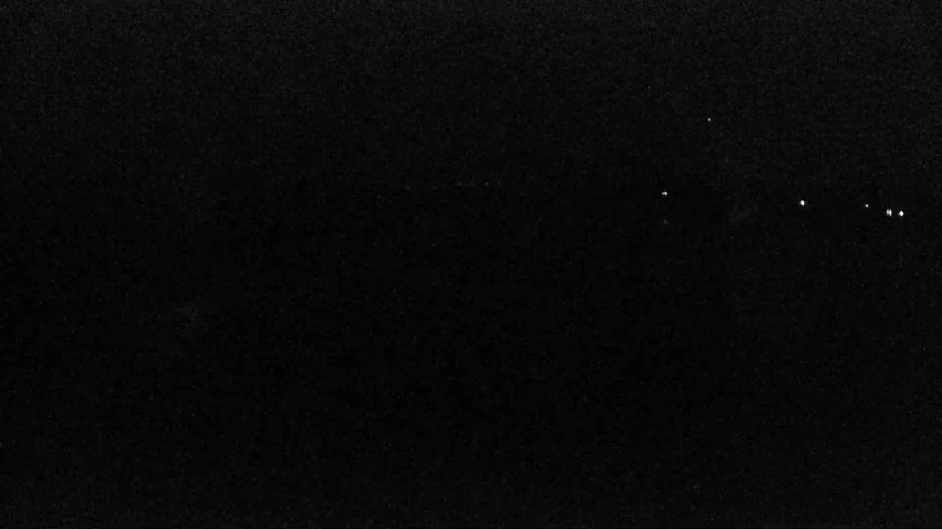 A mostly black image shows a few small, faint white lights in the distance at night.