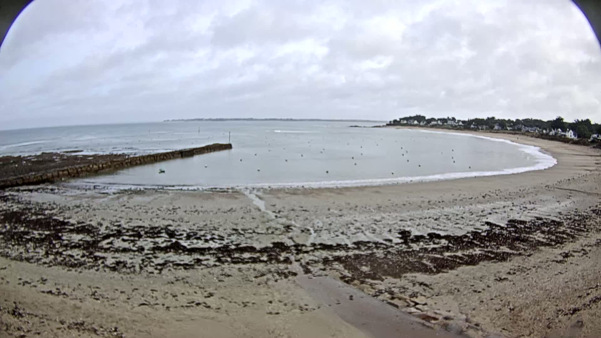 A sandy beach curves around a calm bay under a cloudy sky, with a low stone wall and several small buoys in the water.