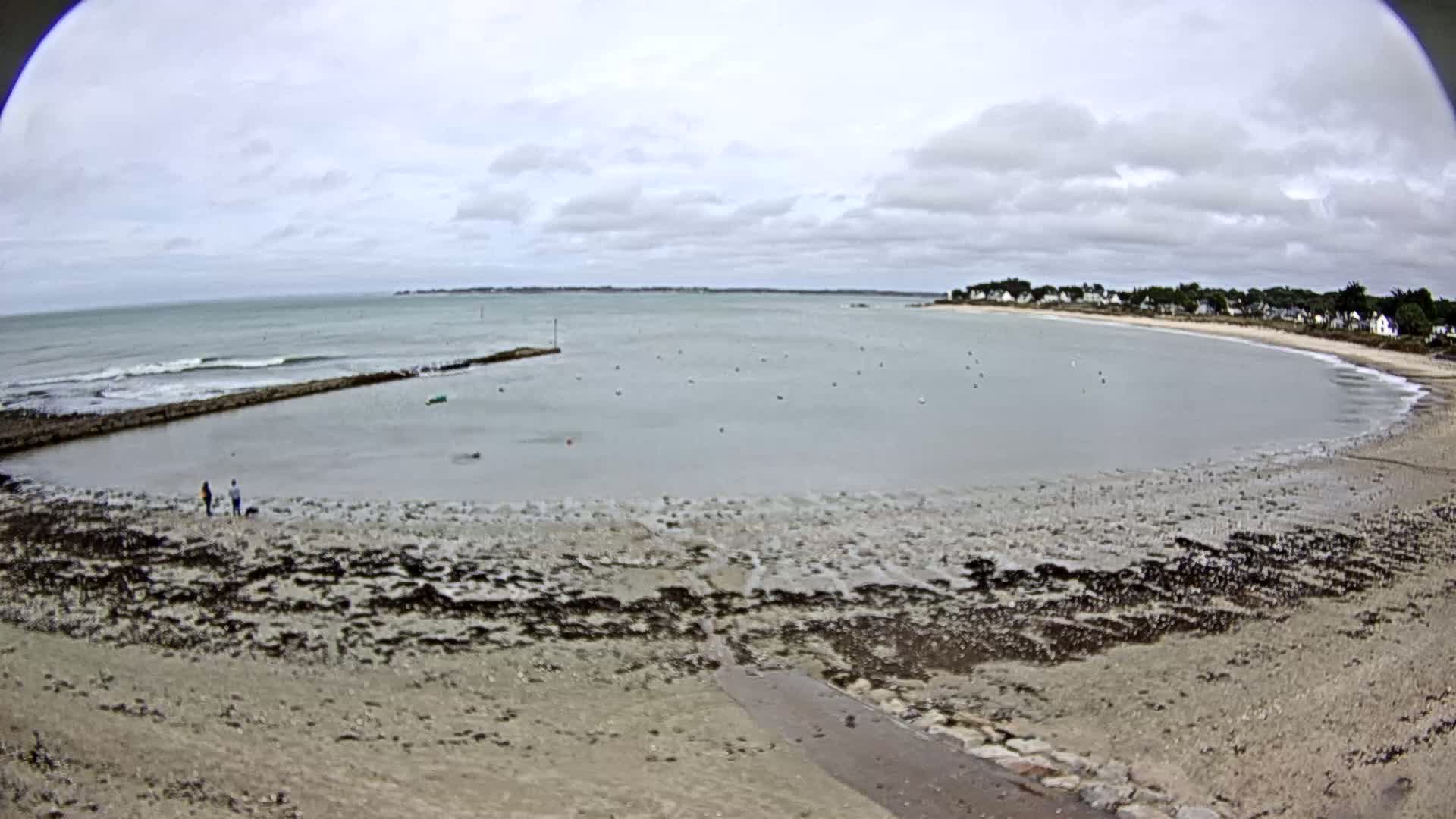 A mostly calm bay with a sandy beach and a few people walking along the shore under an overcast sky.