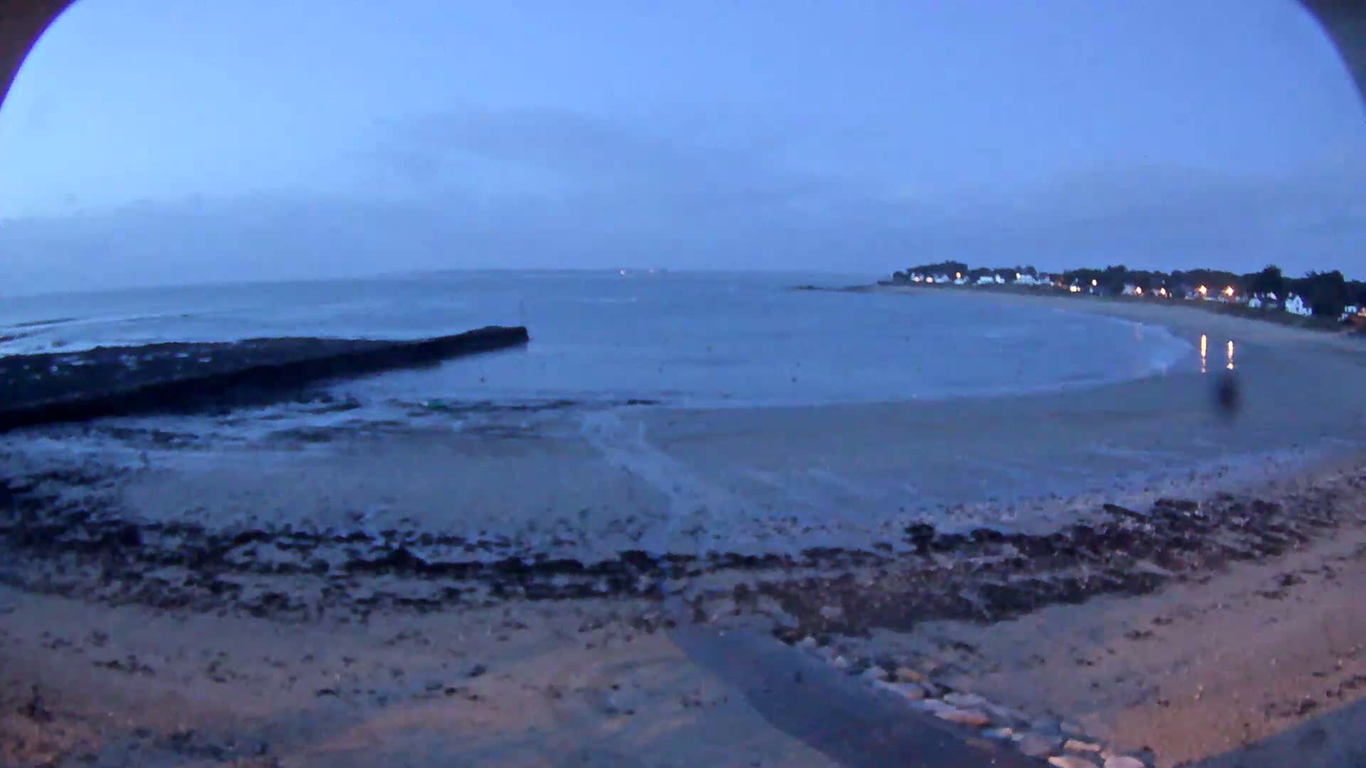 A sandy beach at low tide under a twilight sky, with a calm sea and a small town visible in the distance.