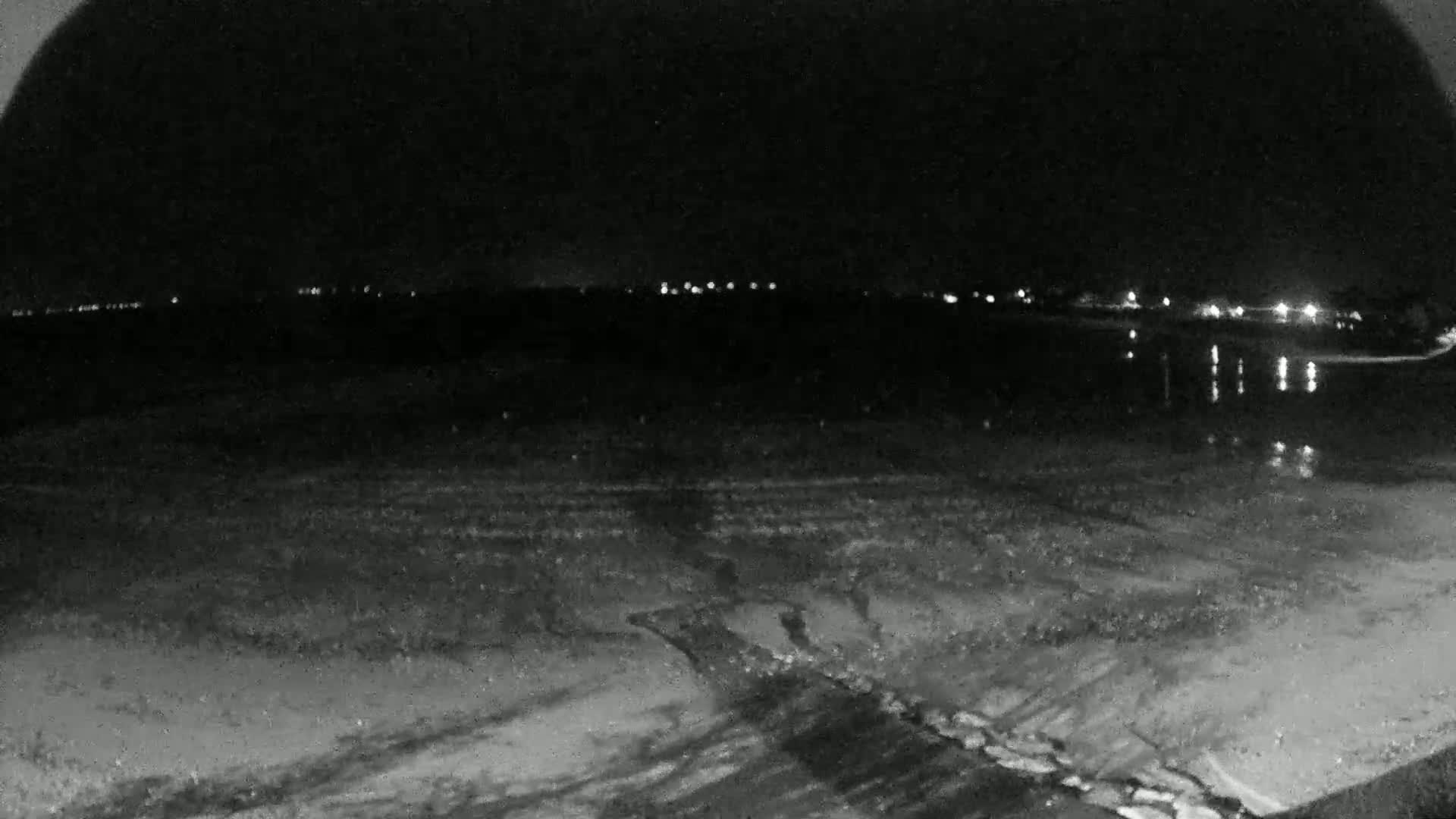 A dark, nighttime view of a beach with calm water reflecting distant lights.