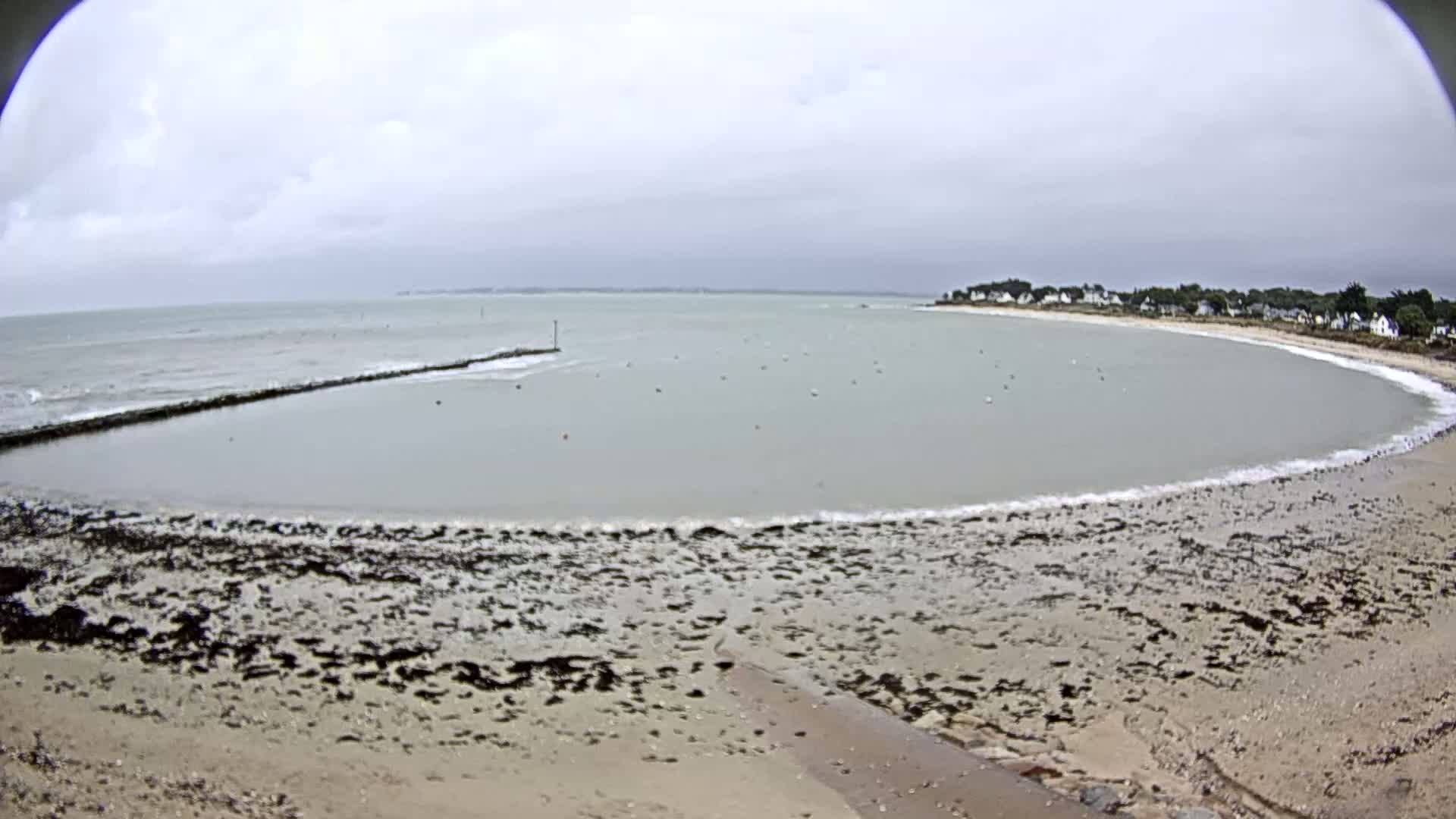 A calm, overcast day at a coastal area shows a sandy beach with dark seaweed patches, a breakwater enclosing a still body of water, and a small town visible in the distance.