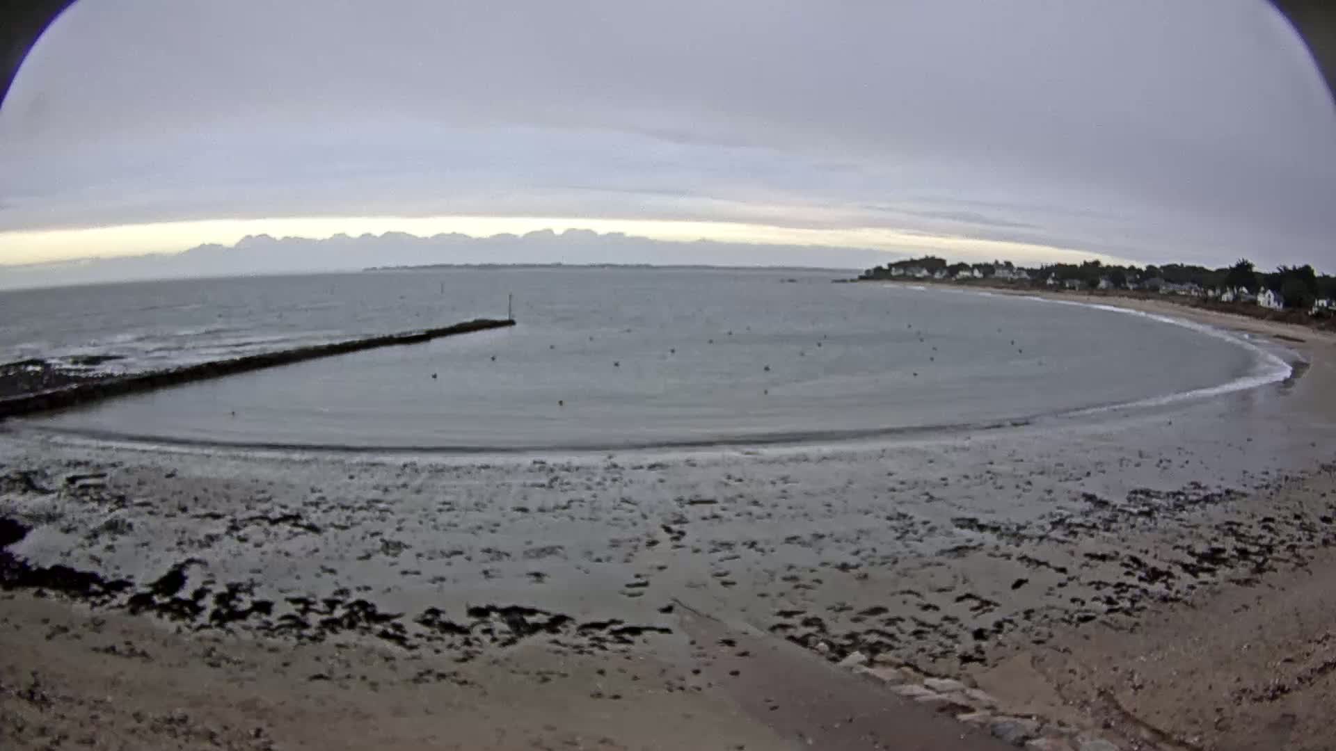 A calm, overcast day reveals a gently curving beach, a small bay with a low stone wall, and distant hills across a body of water.