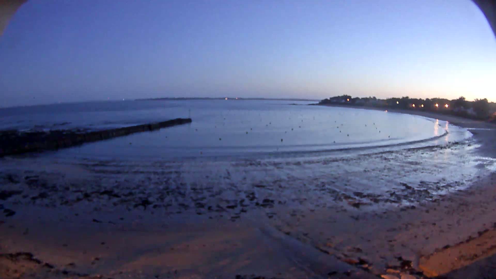 A calm, twilight beach scene shows a gently curving shoreline with a low tide exposing a wide expanse of sand and mudflats, a dark jetty, and a calm bay with small buoys.
