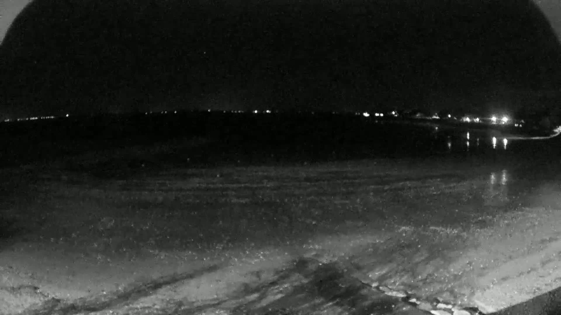 A nighttime, low-light view of a dark, flat expanse of water reflecting distant lights, with a dark shoreline in the foreground.