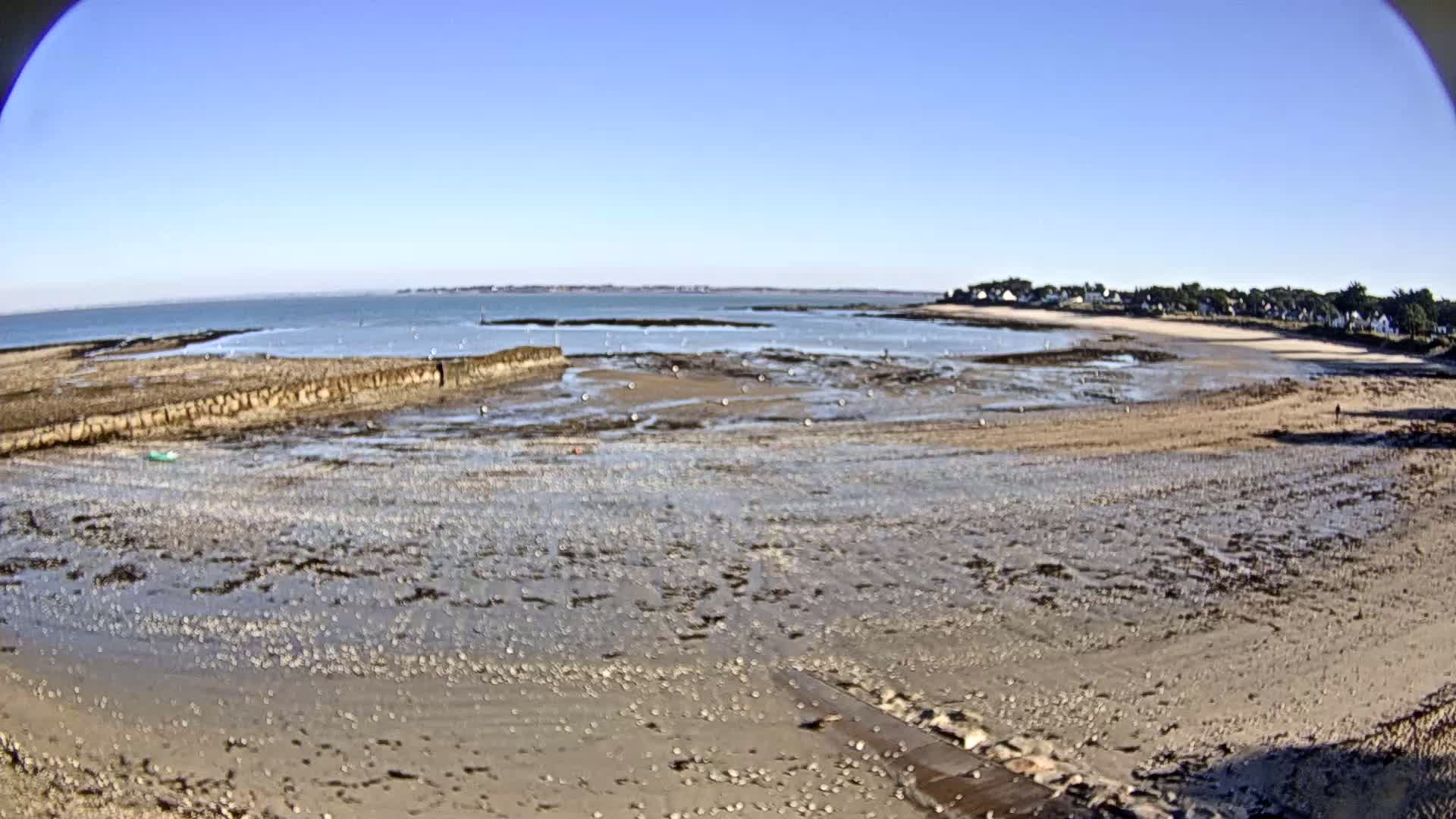 A wide, sandy beach stretches out to a calm sea under a clear blue sky, with low tide exposing rocky intertidal areas and a small cluster of buildings in the distance.
