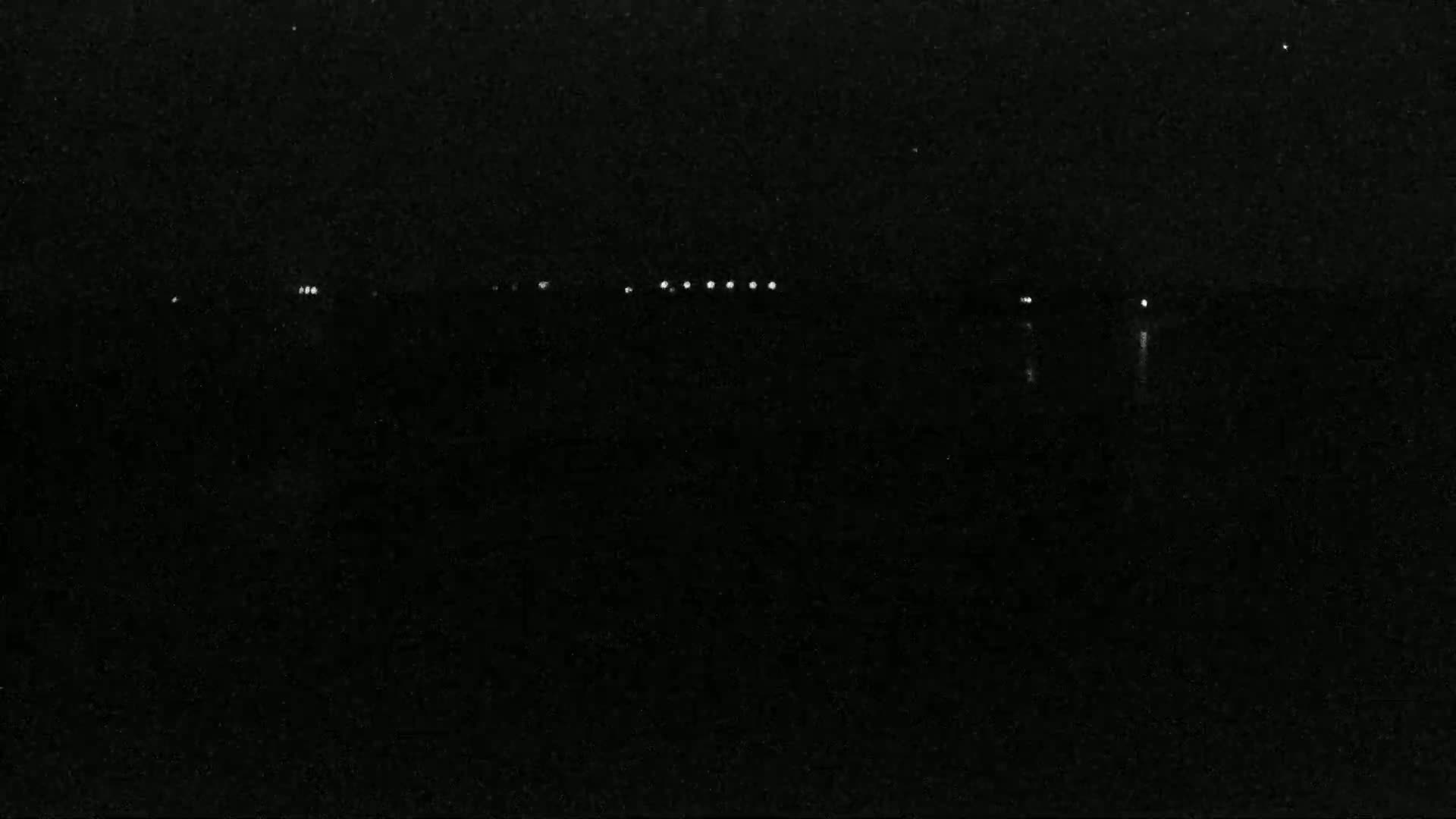 A dark, nighttime scene shows a line of faint lights on a dark body of water.
