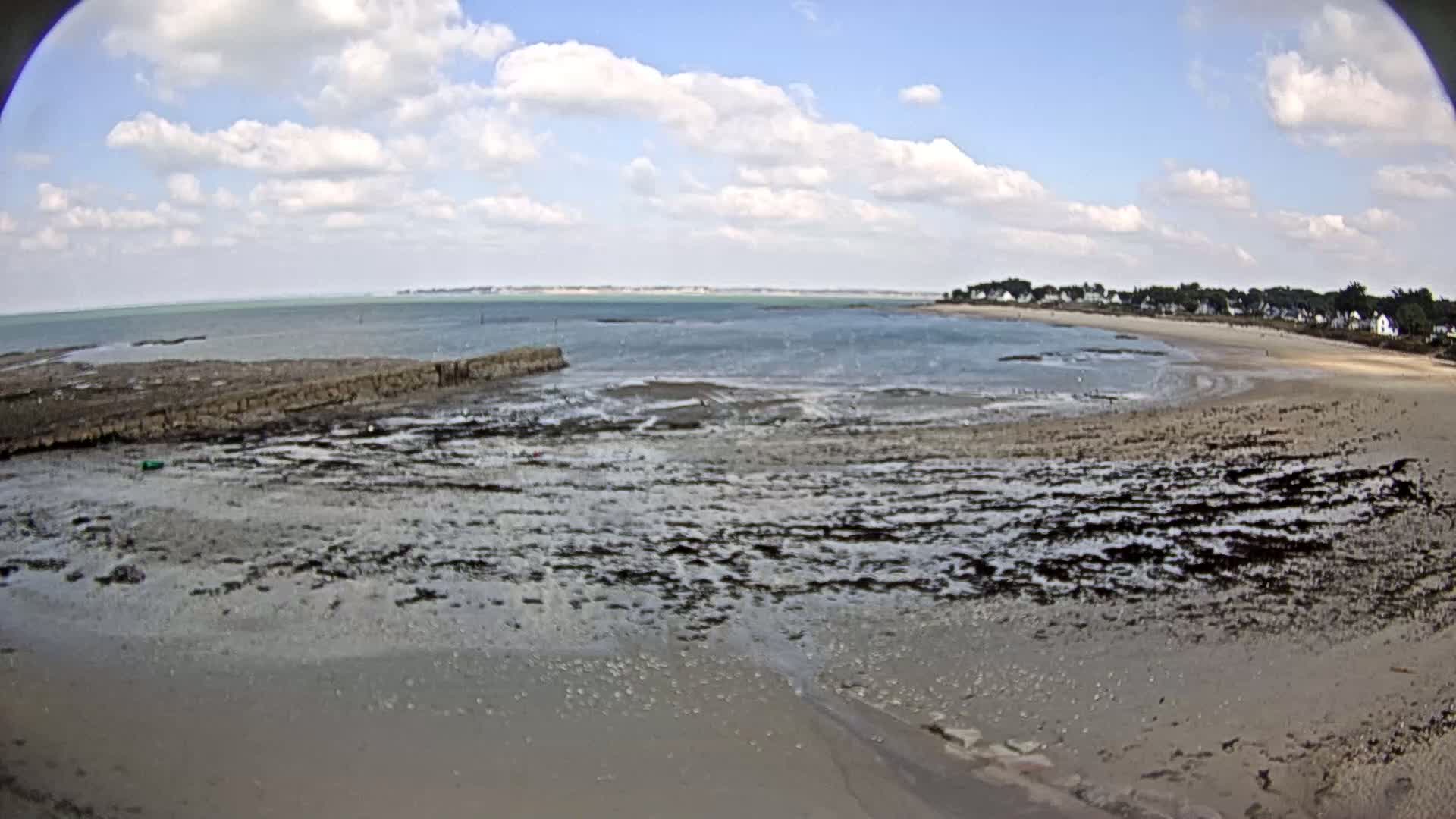 A mostly sunny day reveals a wide, sandy beach with dark, wet rocks exposed at low tide, extending to a calm body of water under a partly cloudy sky.