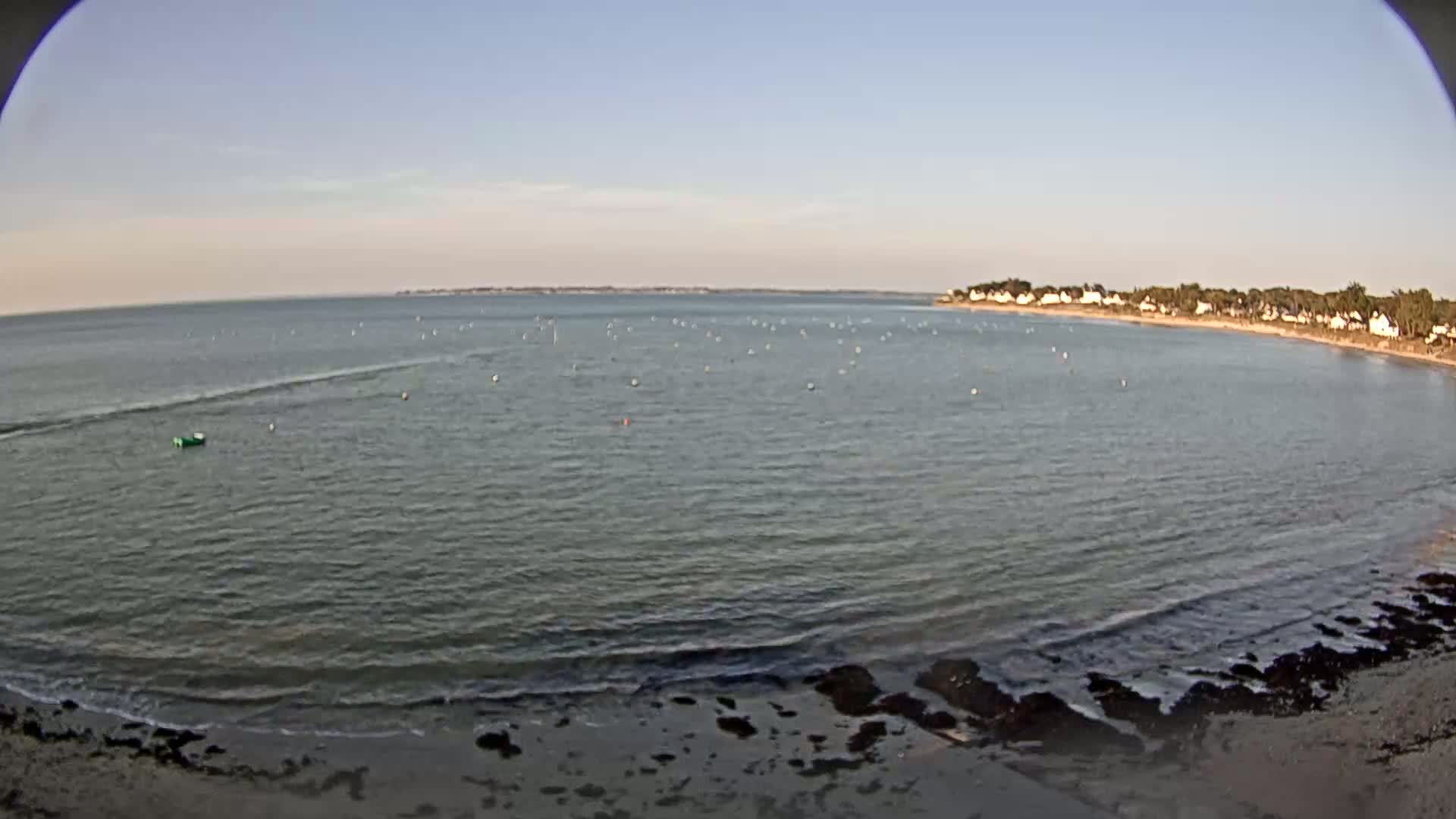 A calm, clear day shows a bay with a sandy shore, small waves, a few boats, and houses lining the opposite shore.