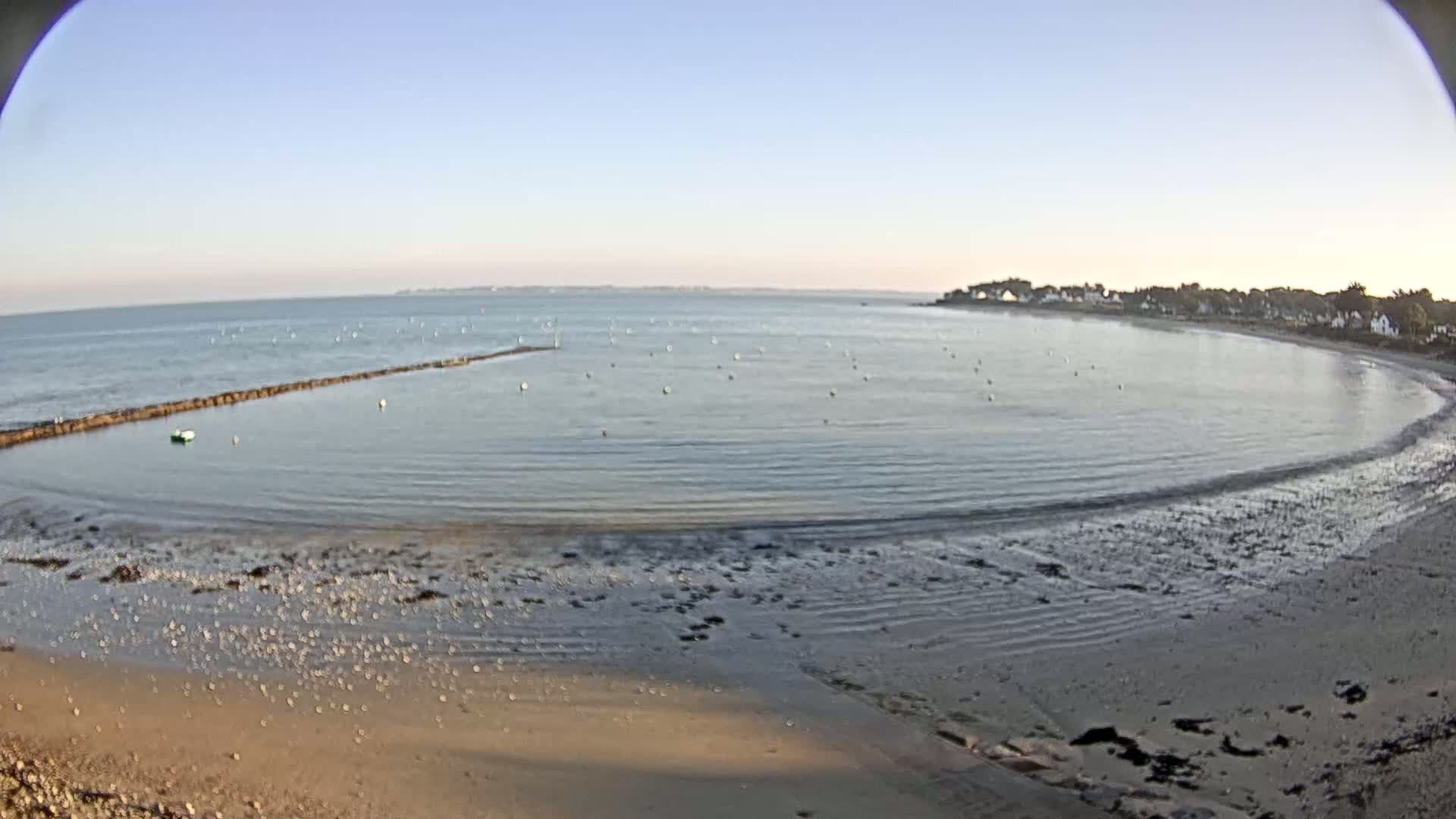 A calm, clear day shows a sandy beach curving around a bay with a low tide revealing a wide expanse of wet sand, a low stone wall partially submerged in the water, and a few small boats.