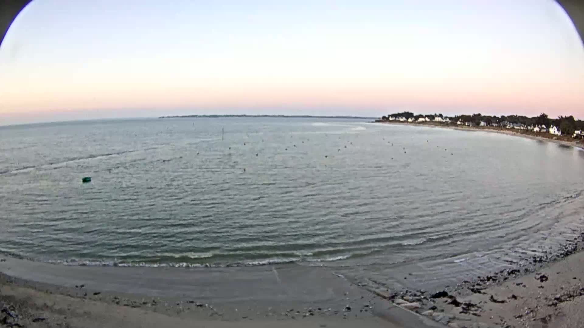 A calm bay with a sandy beach and houses visible in the distance under a pale pink and dusky sky.