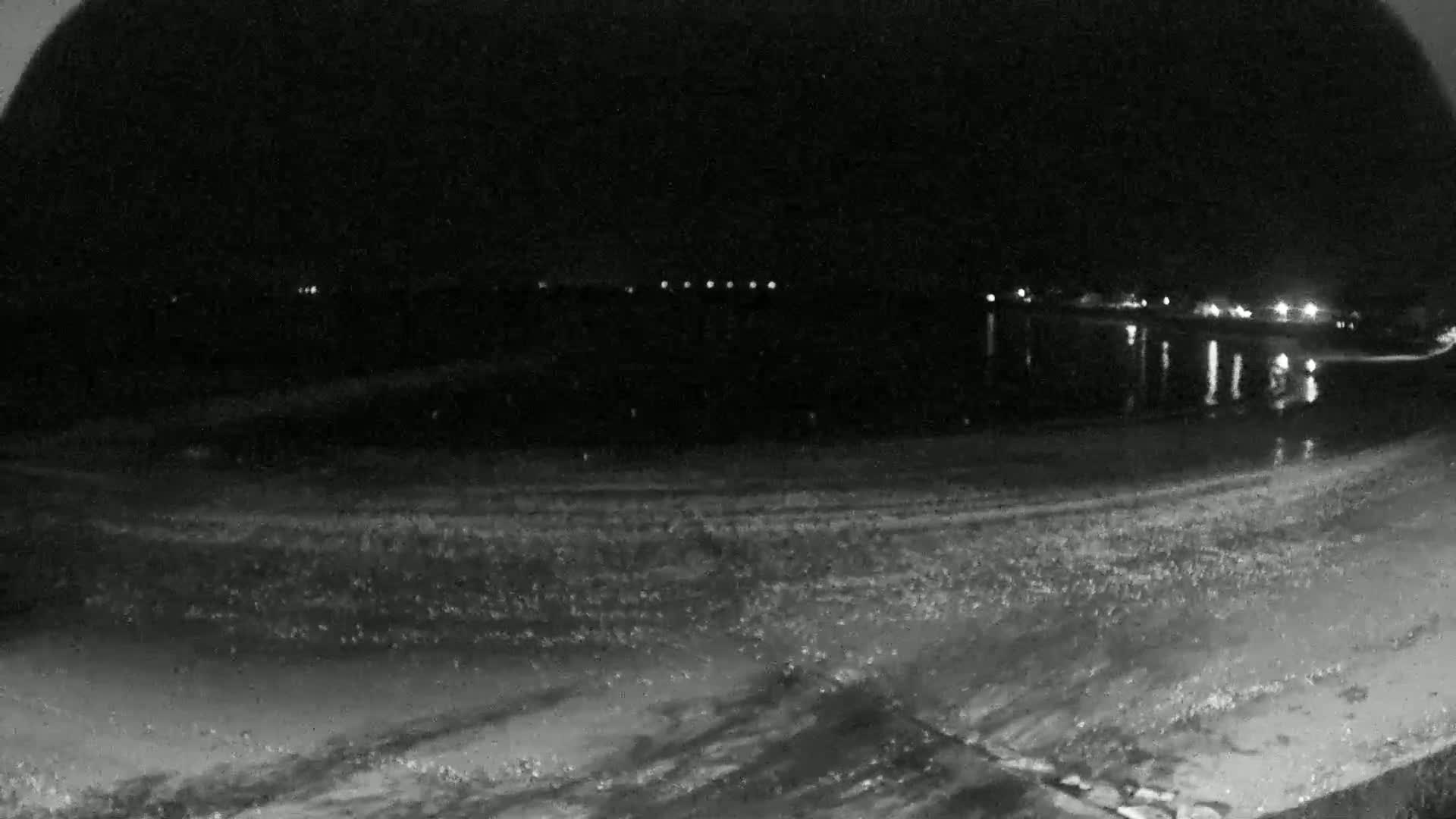 A nighttime view of a dark, sandy shoreline with calm water reflecting distant lights.
