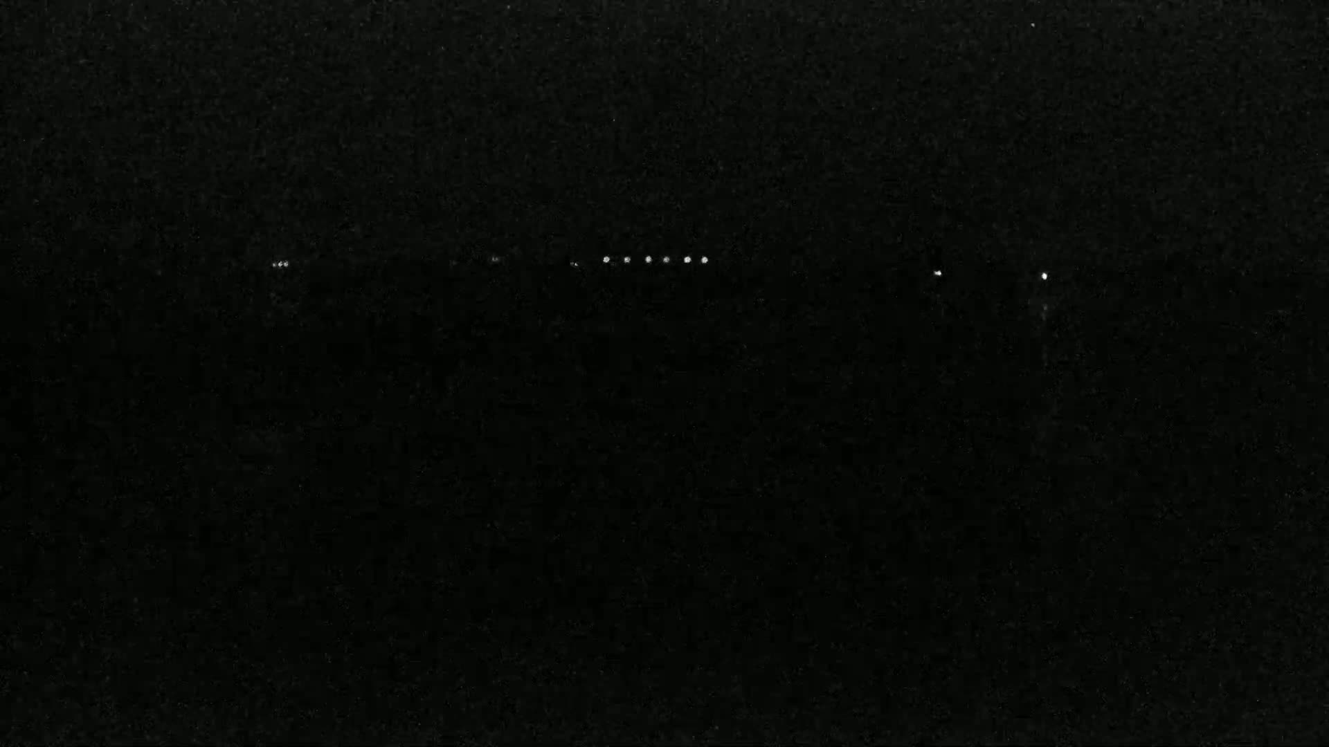 A dark, nighttime scene shows a line of faint lights in the distance.