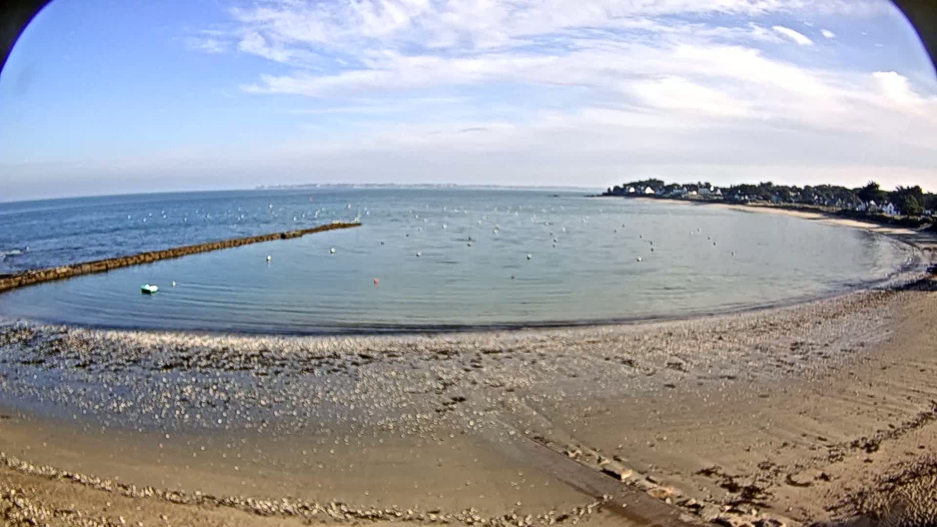 A sandy beach curves around a calm bay under a partly cloudy sky, with a distant shoreline visible across the water.