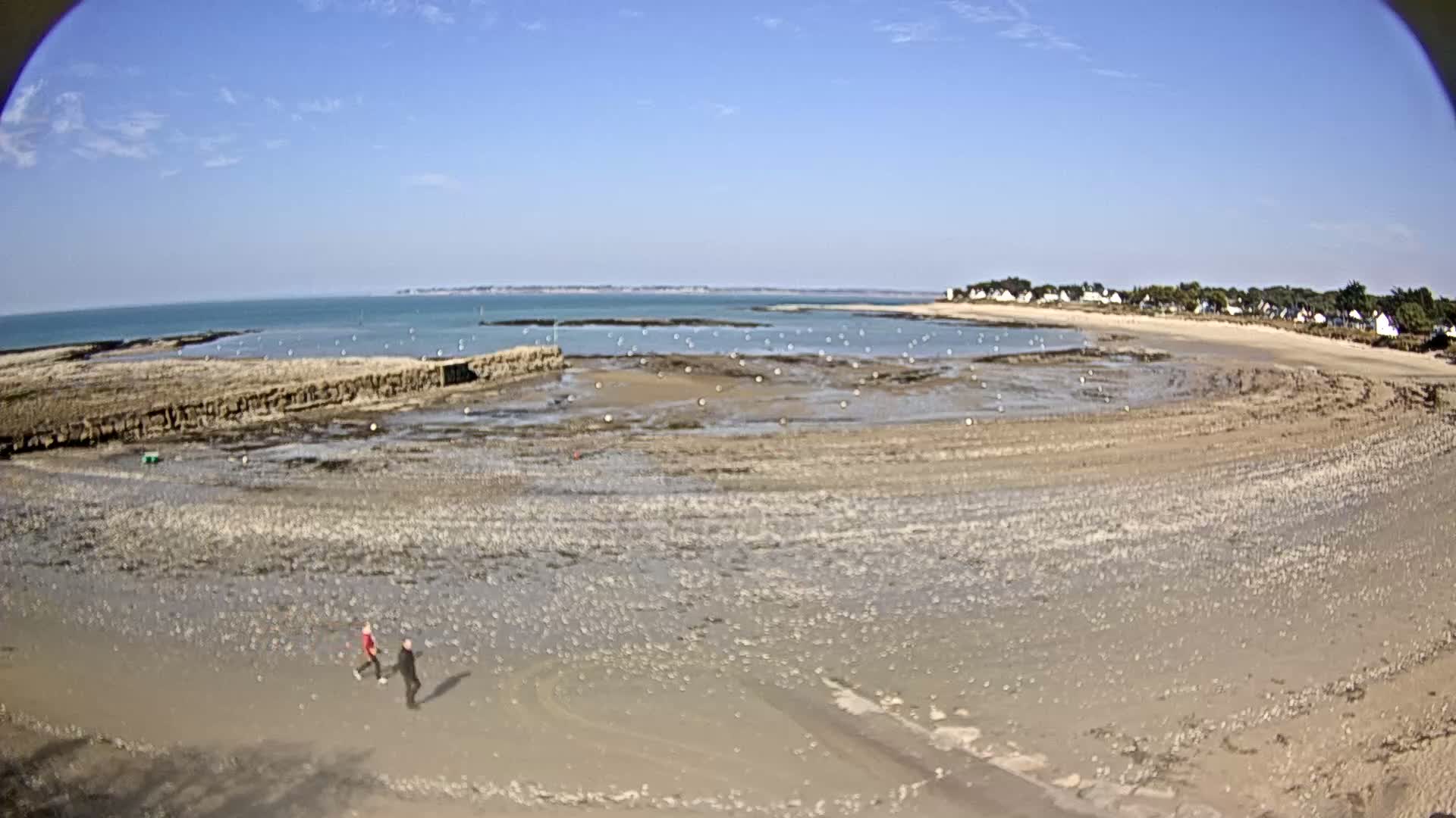 Two people walk on a wide, sandy beach at low tide under a clear blue sky, with a calm sea in the background and a small village visible on the shore.