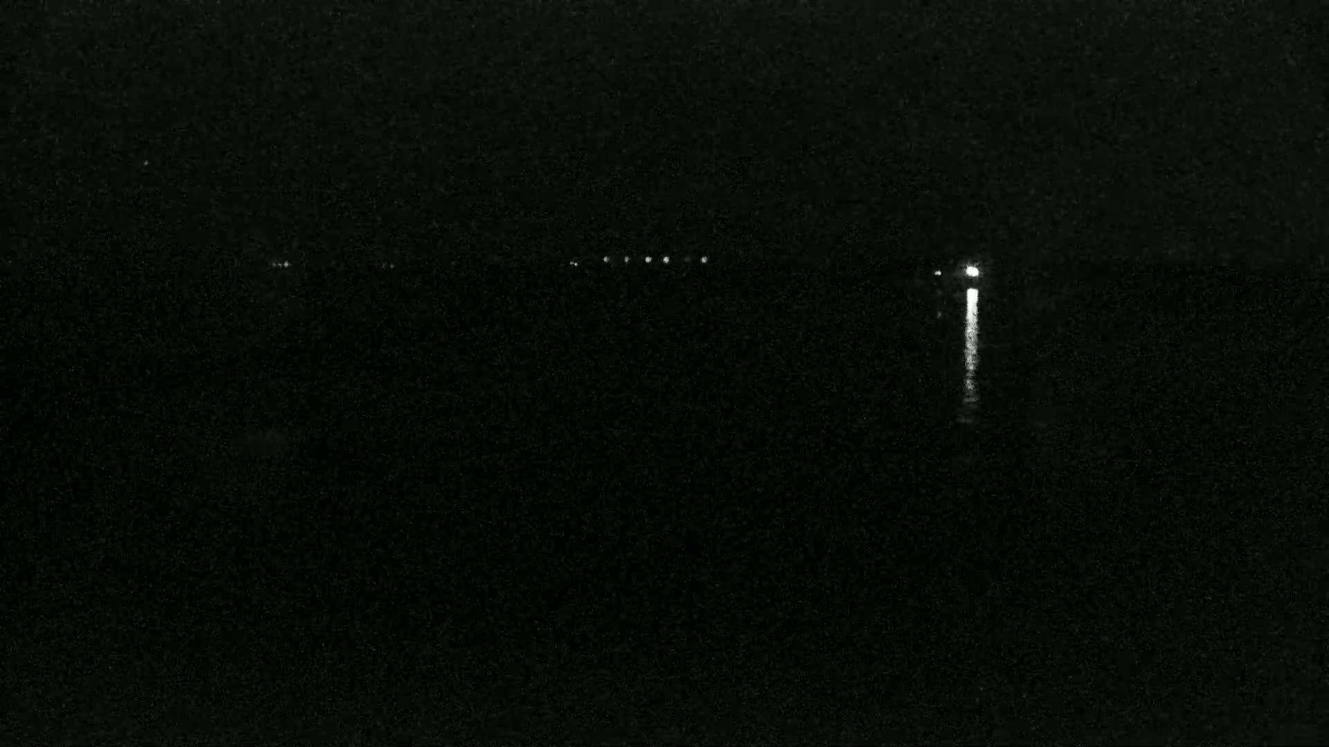 A nighttime view of a dark body of water with a few faint lights visible on the horizon and a brighter light reflecting on the water in the distance.